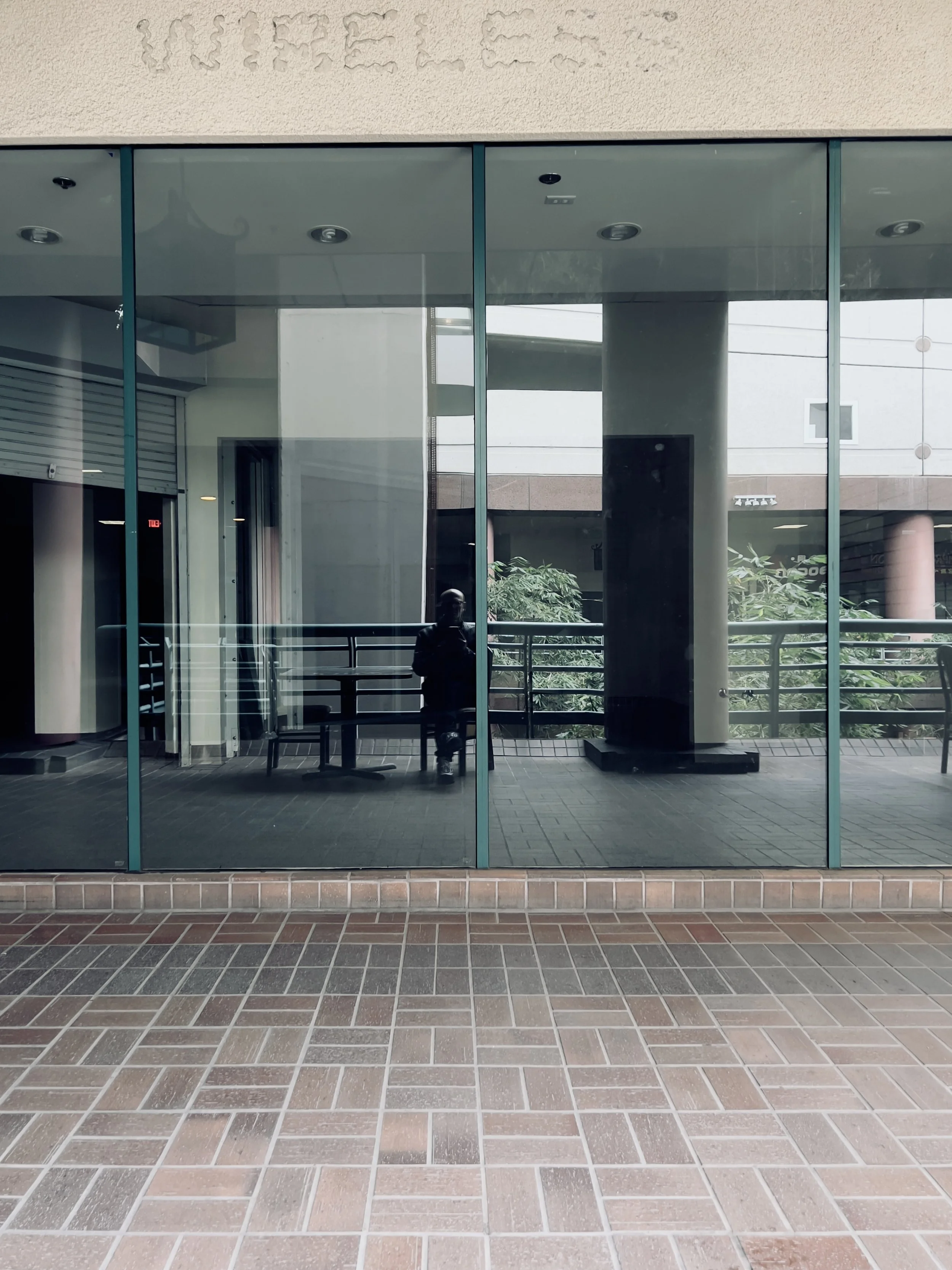 Reflections on large glass windows showing a person sitting, brick pavement, and interior of a building.