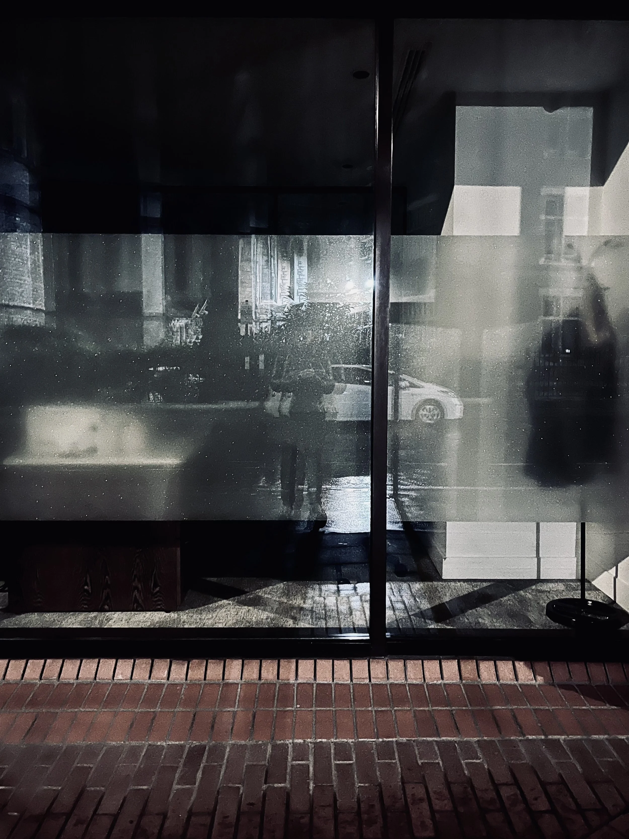 Reflection of a person taking a photo in glass window on a city street, with cars and buildings visible outside.
