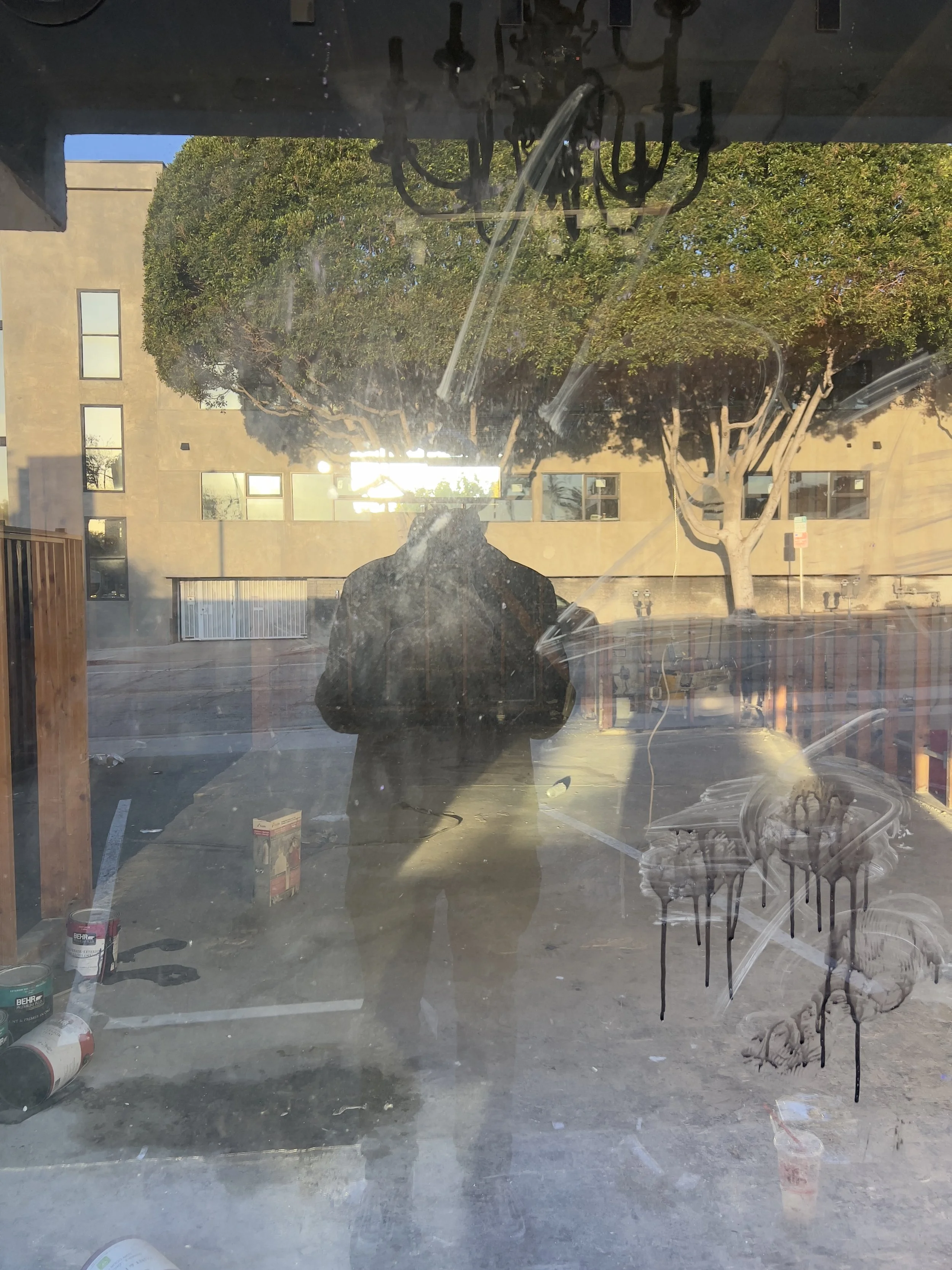 Reflection of a person taking a photo in a dirty glass window with smudges and paint drips, outside view of trees, buildings, and parking lot.