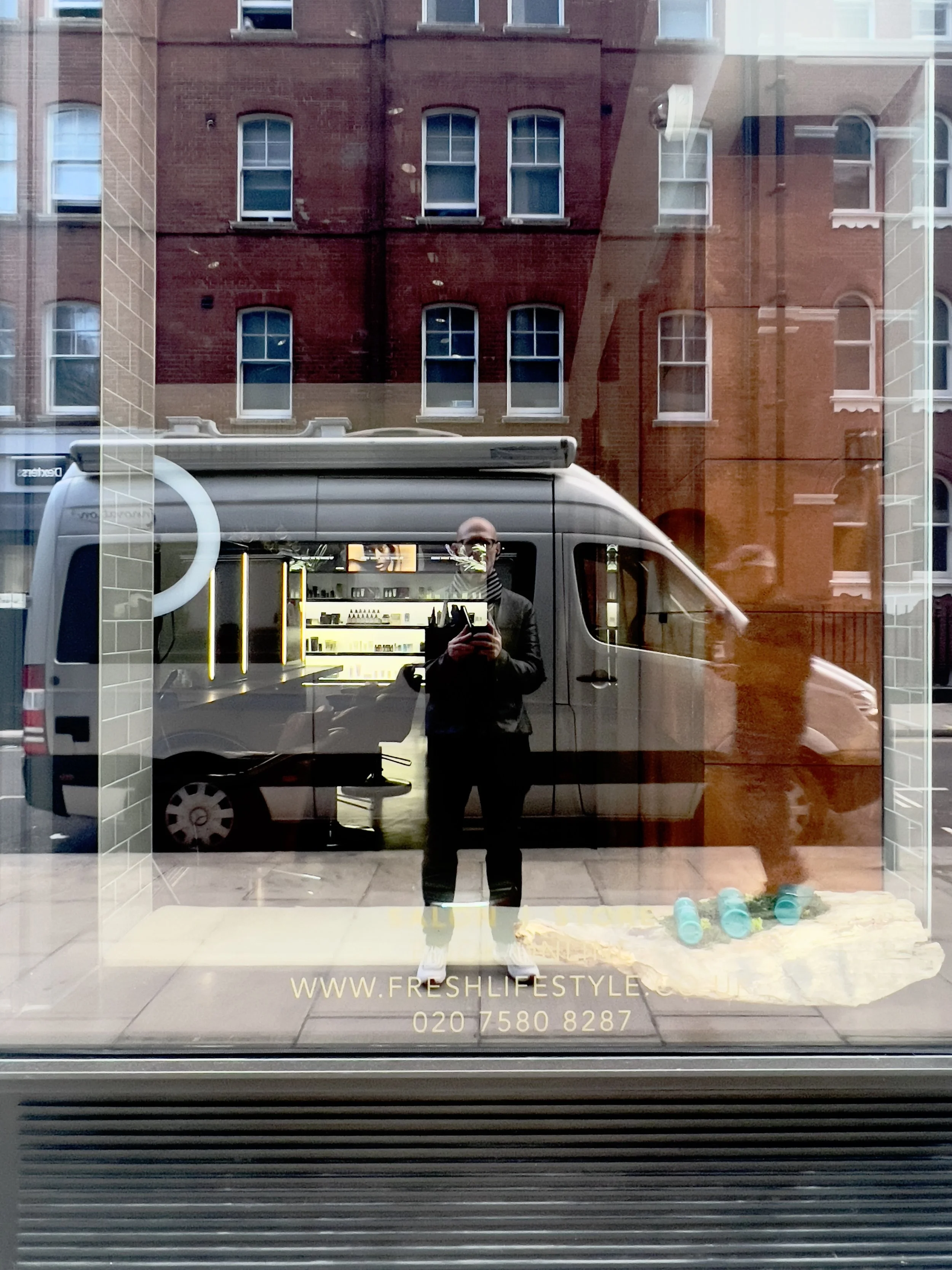 Reflection of a street with a person taking a photo, a van, and a brick building in the background, seen through a shop window displaying hair care products. The window has "www.freshlifestyle.co.uk" and a phone number written on it.