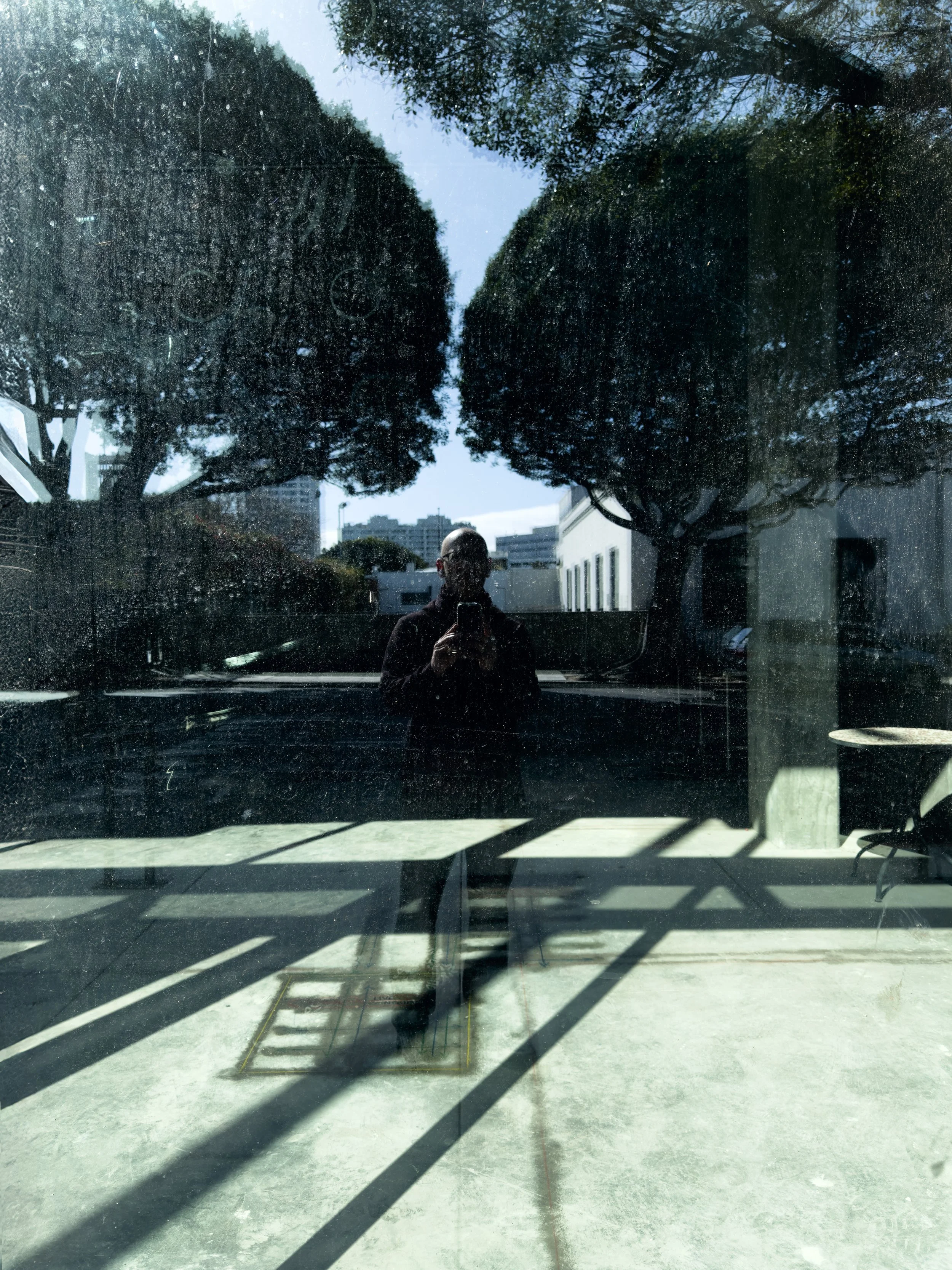 Reflection of a person taking a photo with a phone, seen through a dirty glass window, with trees and buildings in the background.