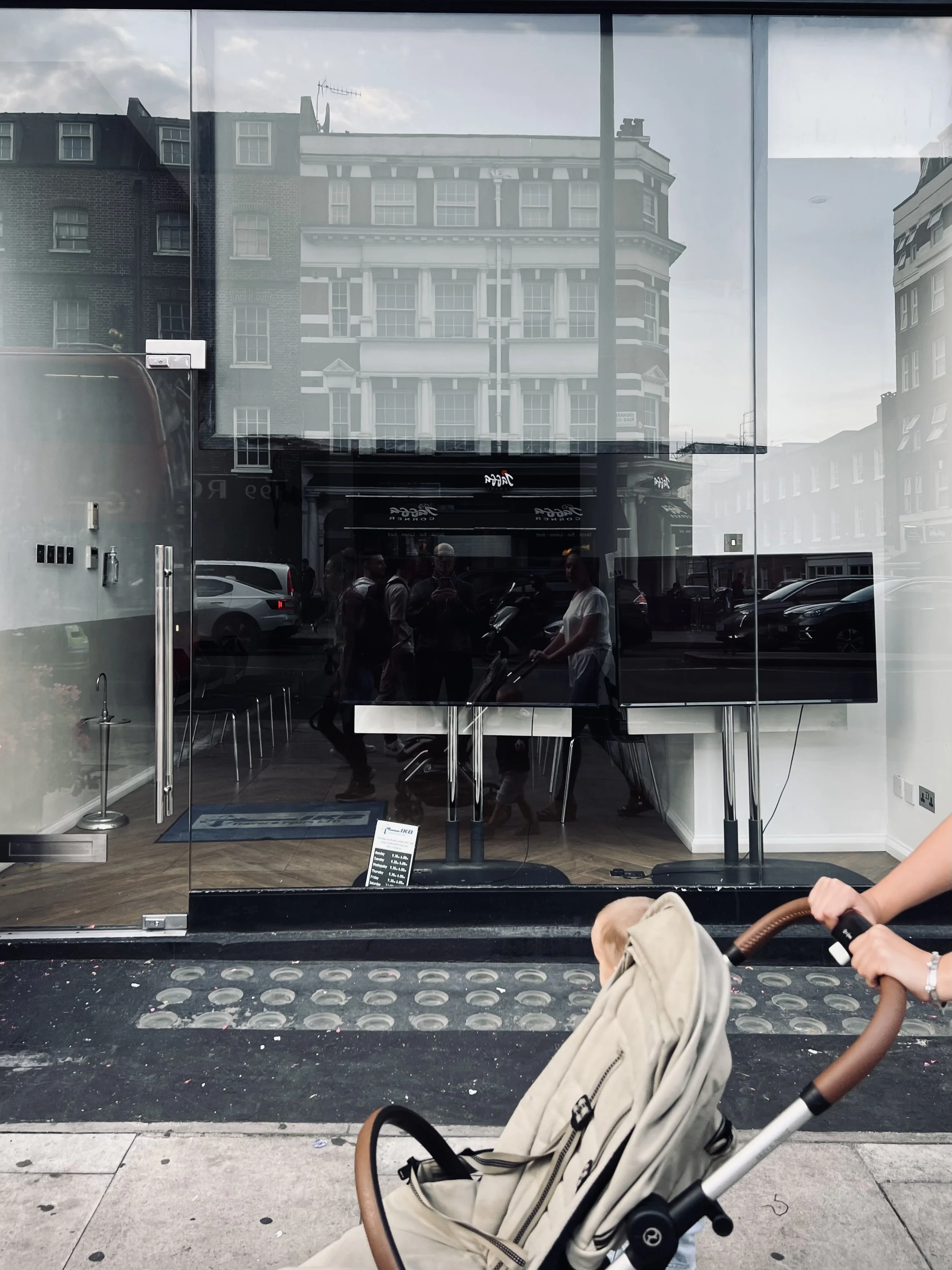 City sidewalk scene with a glass storefront reflecting buildings and people walking, with part of a person pushing a stroller in the foreground.