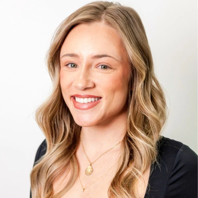 A young woman with long, wavy blonde hair, smiling, dressed in a black top, wearing a gold necklace, against a plain light background.