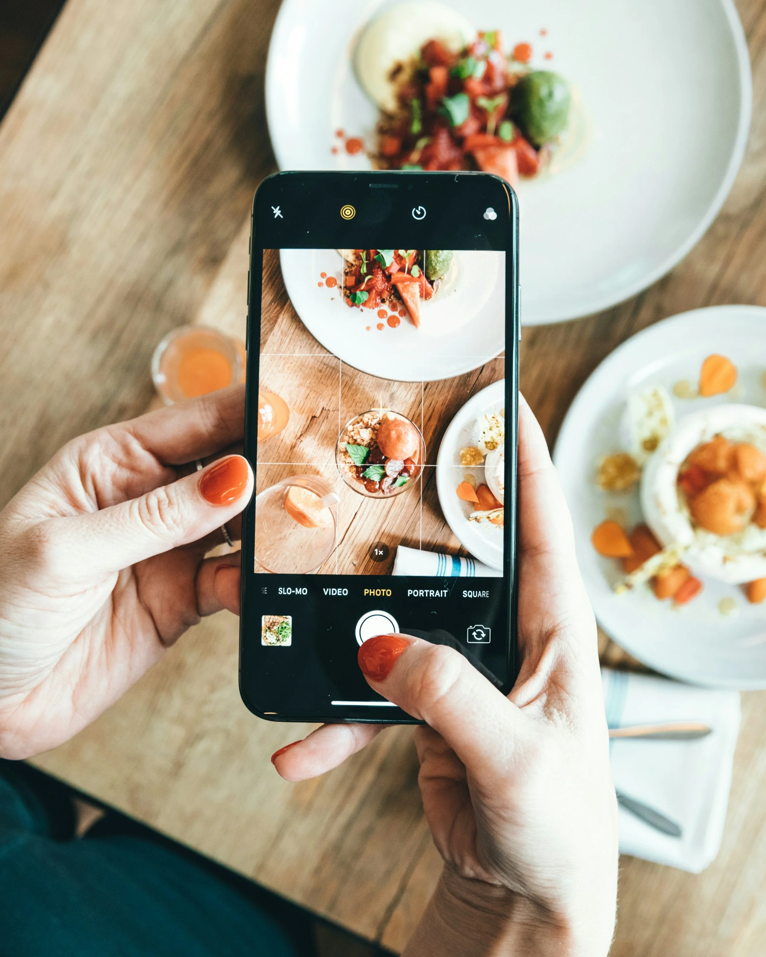 Person taking a photo of plated food with smartphone, focus on food and camera screen, desserts with sauces and garnishes on white plates, wooden table, drink in background.