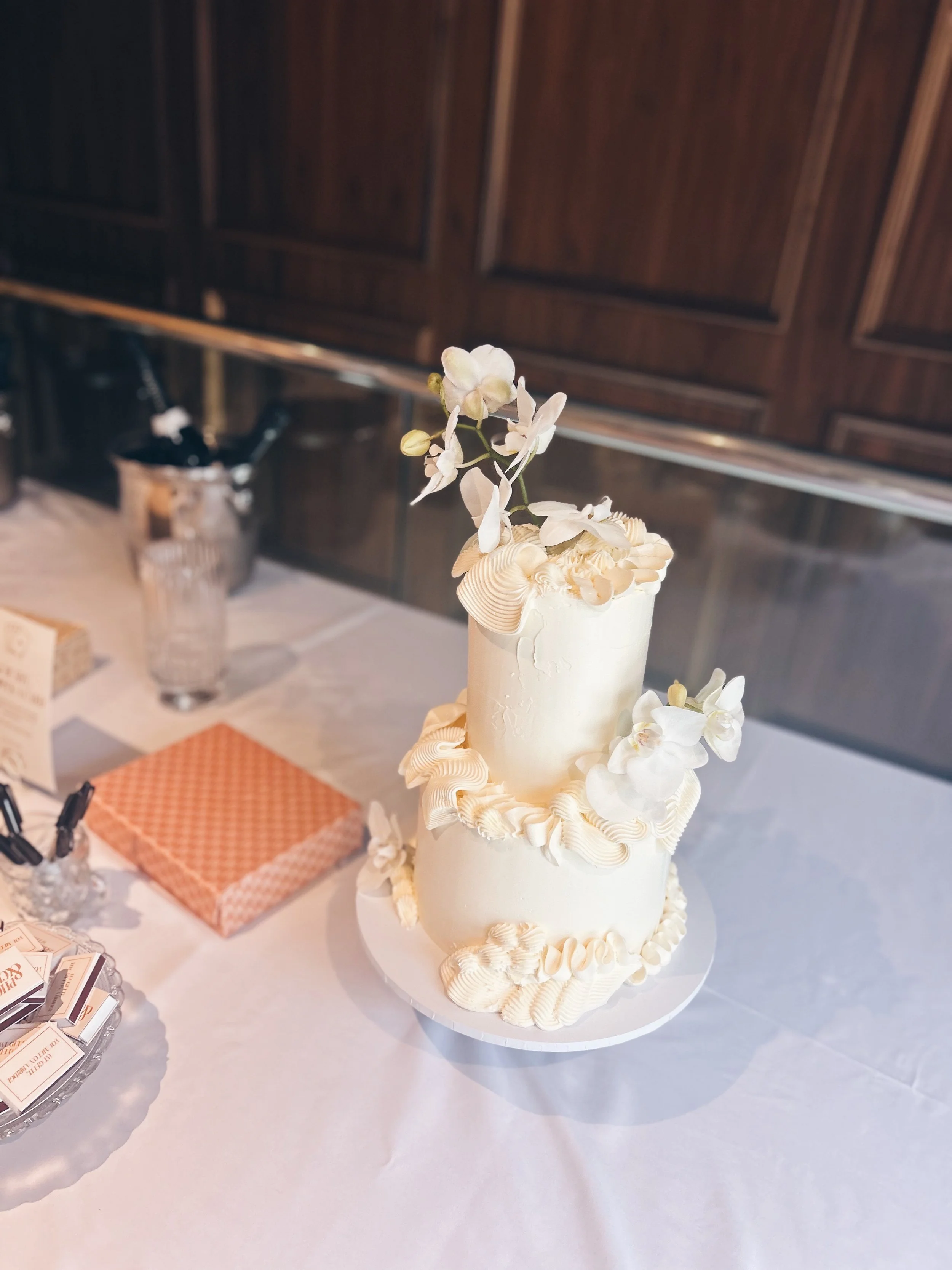 Elegant two-tier white wedding cake decorated with white orchid flowers and cream-colored piped icing, placed on a white tablecloth.