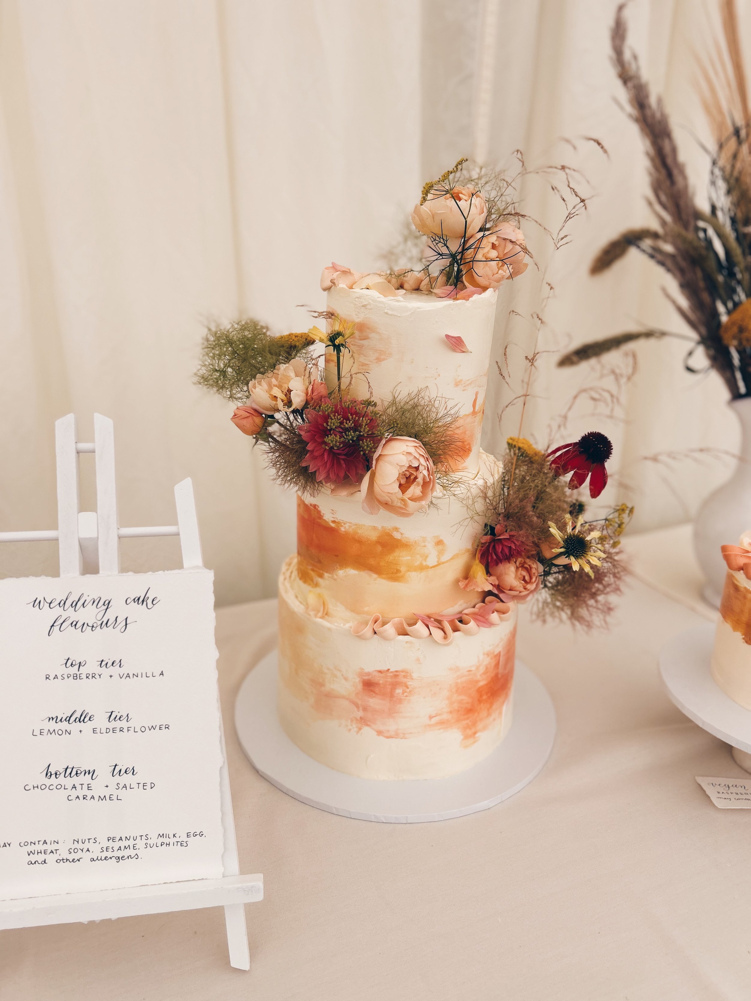 A three-tier wedding cake with a white and peach marble design, decorated with pink and red flowers, placed on a white table with a wedding cake flavor sign beside it.