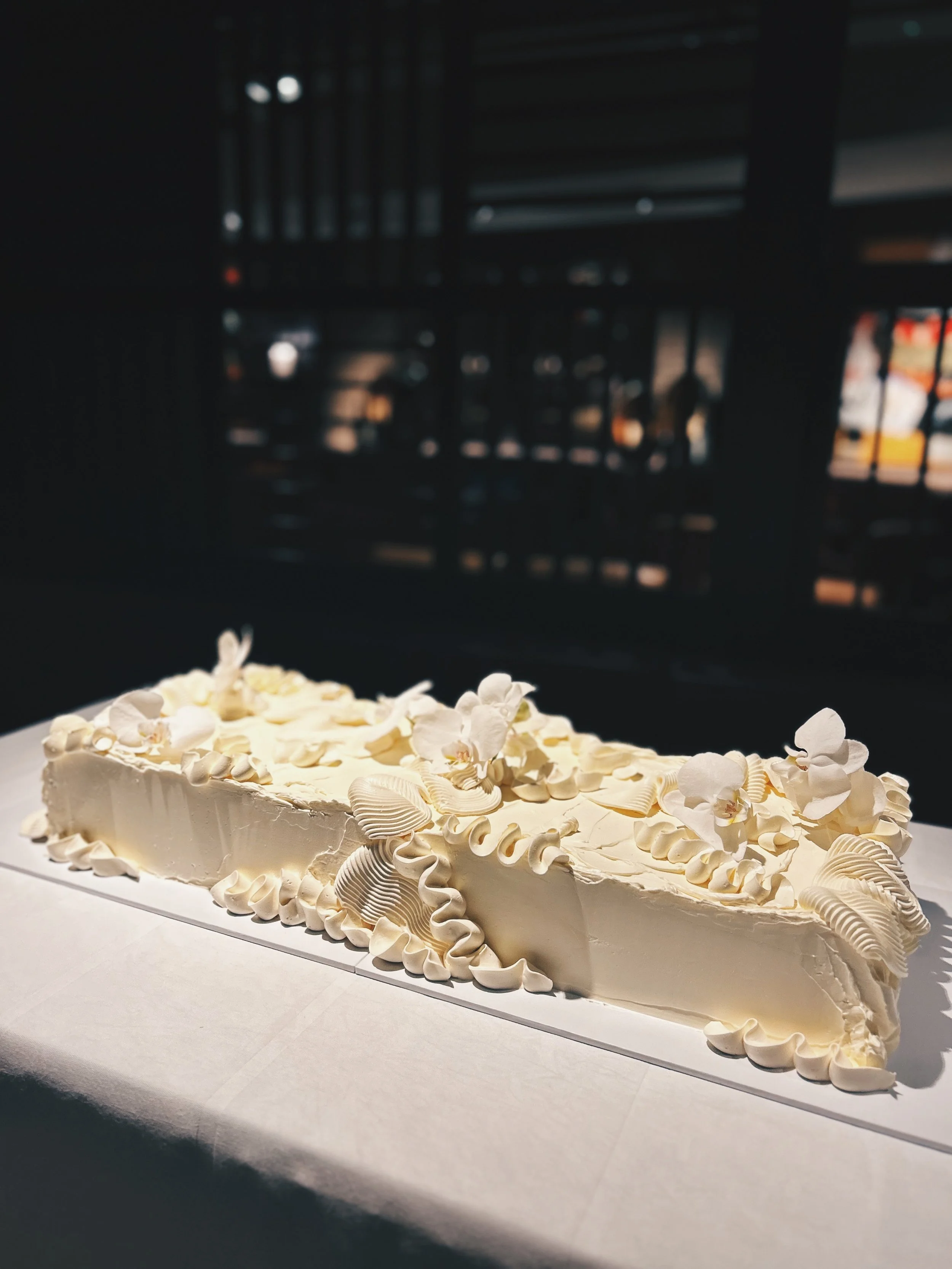A rectangular, white frosted cake decorated with white flower and shell-shaped icing details, placed on a white tablecloth with a dark background.