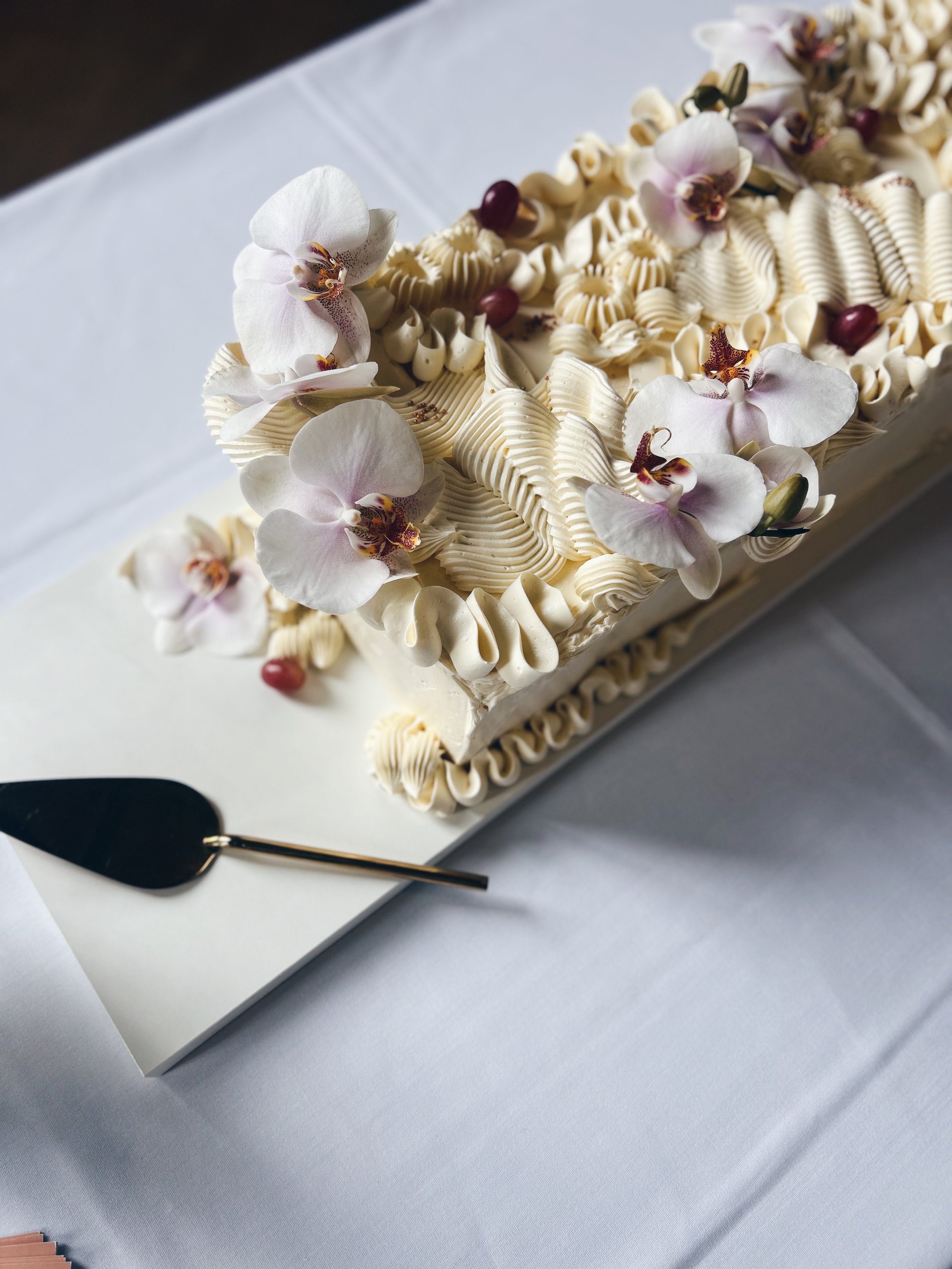 A rectangular wedding cake decorated with white frosting, pink orchids, and grapes, placed on a white surface with a cake server nearby.