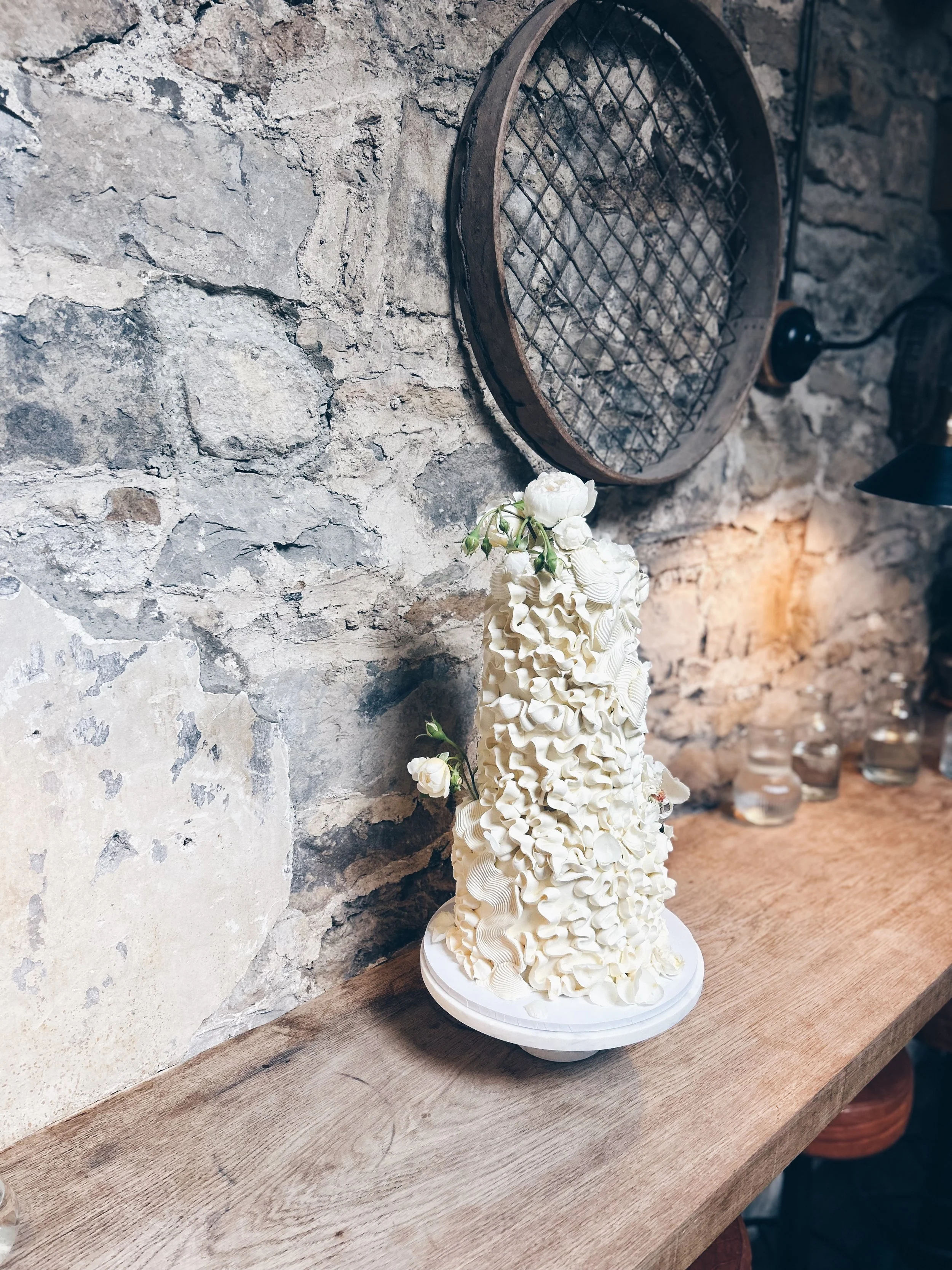 A tall, white, ruffled cake decorated with small white flowers on a white cake stand, placed on a wooden surface against a stone wall background.