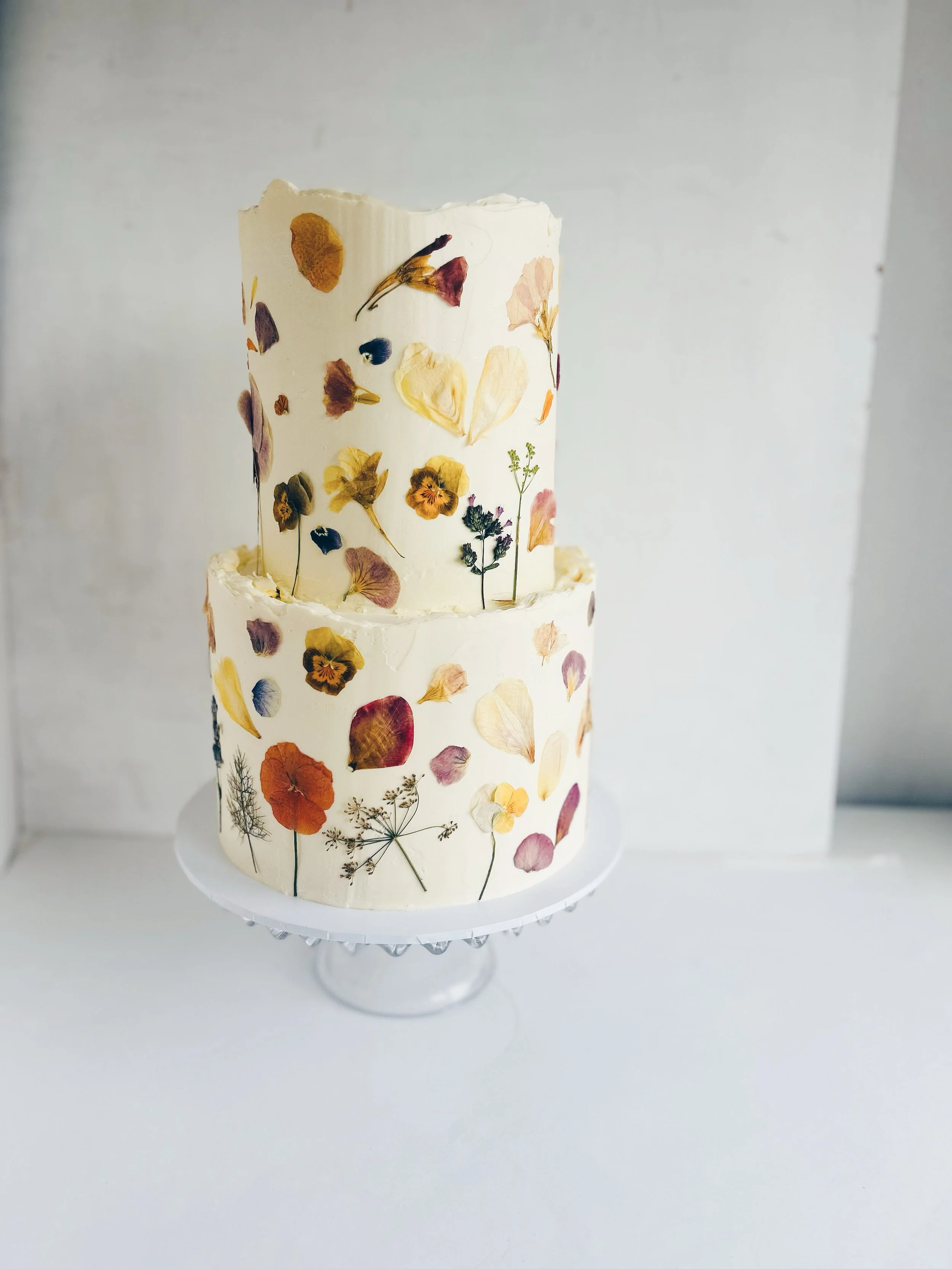 A two-tiered cake decorated with pressed flower petals, displayed on a clear glass cake stand against a plain, light-colored background.