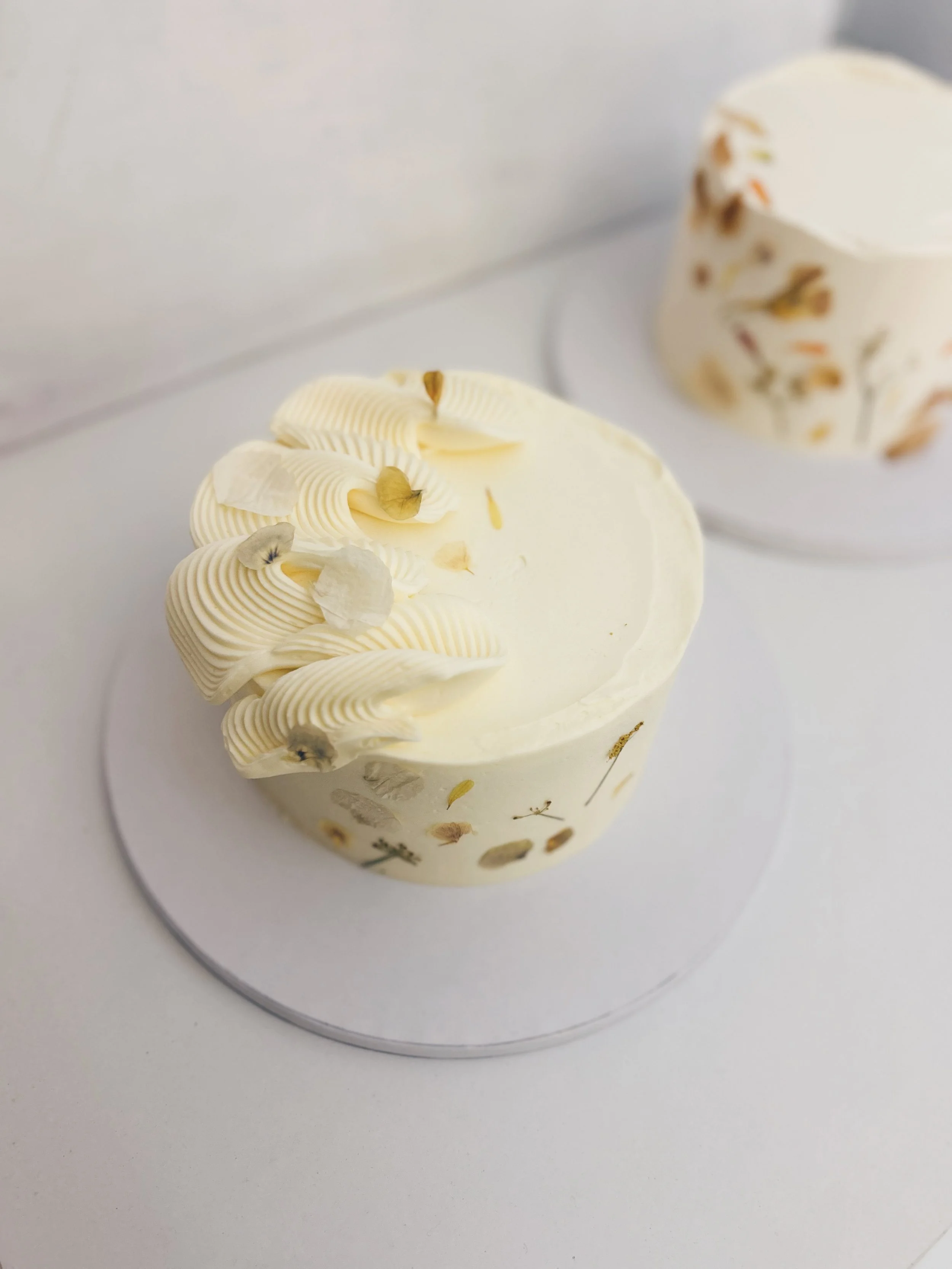 A cake with white frosting decorated with piped cream and edible flower petals, placed on a white cake board.