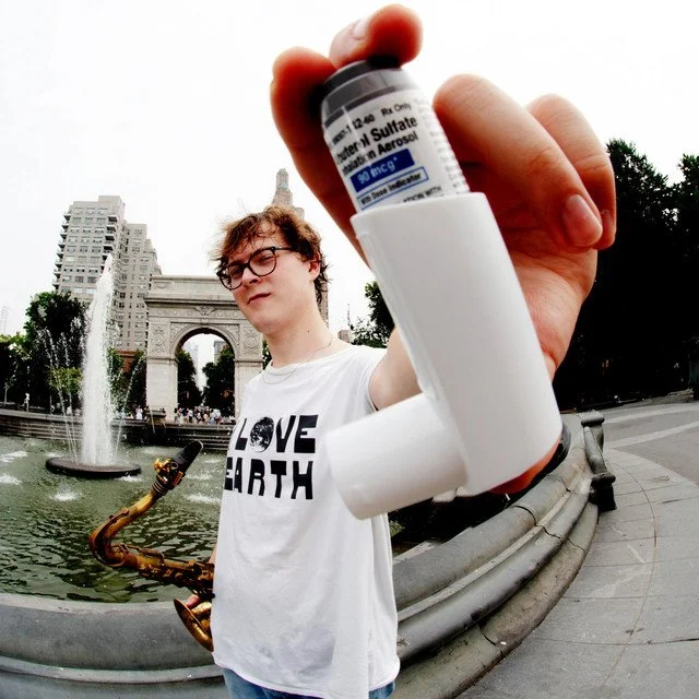 Person holding an asthma inhaler in front of a fountain and cityscape, wearing a white t-shirt with 'LOVE EARTH' printed on it. Album artwork for "Wayward Star" by Ben Lindenburg.