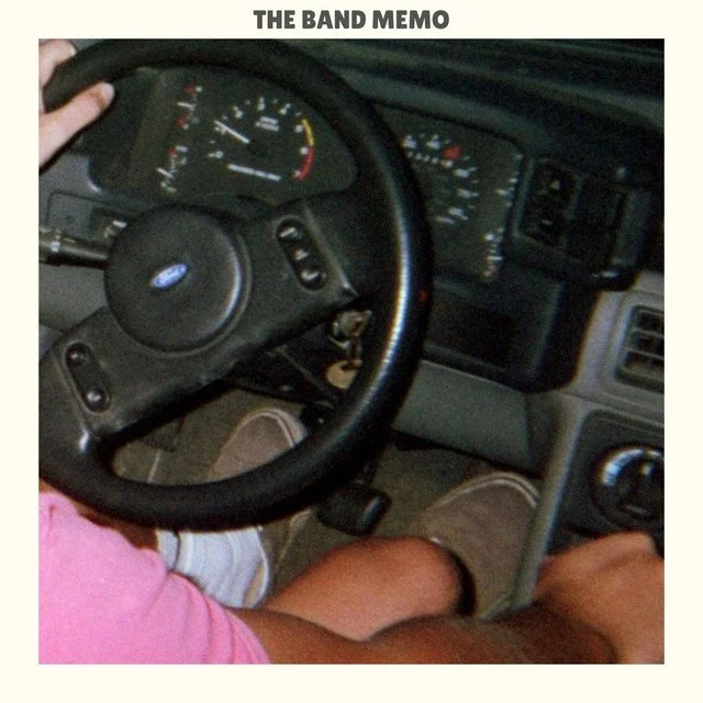Album artwork for "Love on Time" by the band The Band Memo, featuring an inside view of a vehicle dashboard with a steering wheel, speedometer, and other instrument gauges. A person wearing pink and beige pants is seated in the driver's seat.