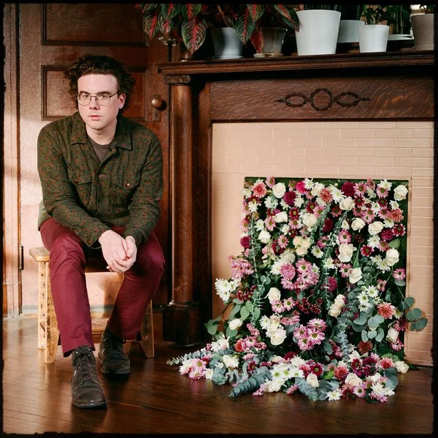 A young man with glasses and dark hair, wearing a dark green patterned shirt and red pants, sits on a wooden chair next to a fireplace. Next to him, there is a large arrangement of pink, white, and purple flowers leaning against the fireplace