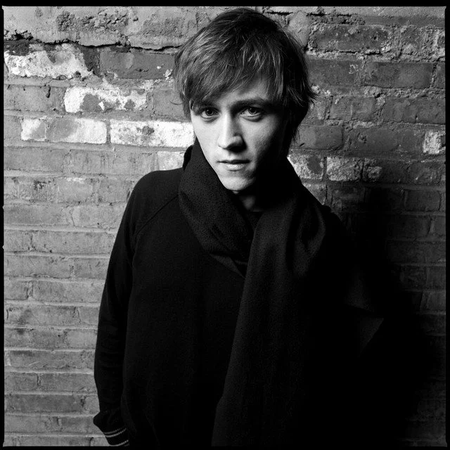 Black and white portrait of a young man with short, tousled hair standing against a brick wall, wearing a dark jacket and scarf. Album artwork for the song "You are Impossible" by Sondre Lerche