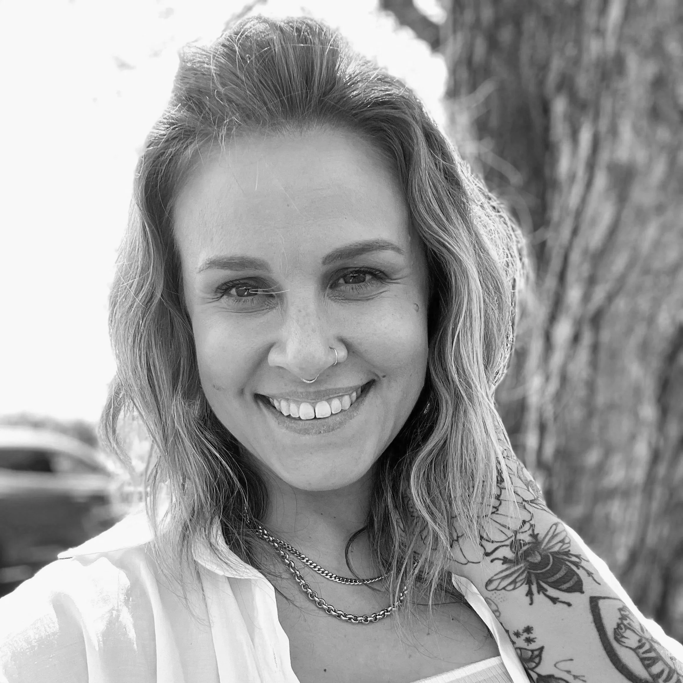 A woman with wavy, shoulder-length hair smiling outdoors with a tree in the background. She has a nose piercing and tattoos on her arm, wears gold necklaces and a white top.