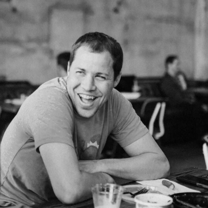 A man in a green t-shirt sitting at a table, smiling and laughing in a restaurant or cafe setting.