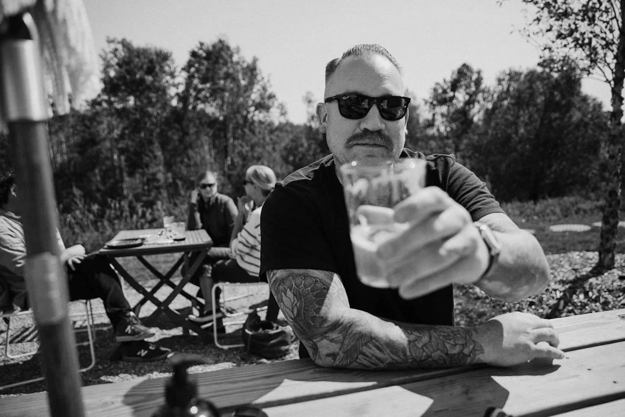 Man with tattoos and sunglasses sitting at a wooden picnic table outdoors, holding a glass of beer toward the camera. Other people are sitting at a table in the background, surrounded by trees.