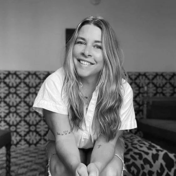 A young woman with long blonde hair and a white shirt, smiling at the camera in a cozy, warmly lit room with patterned seating.