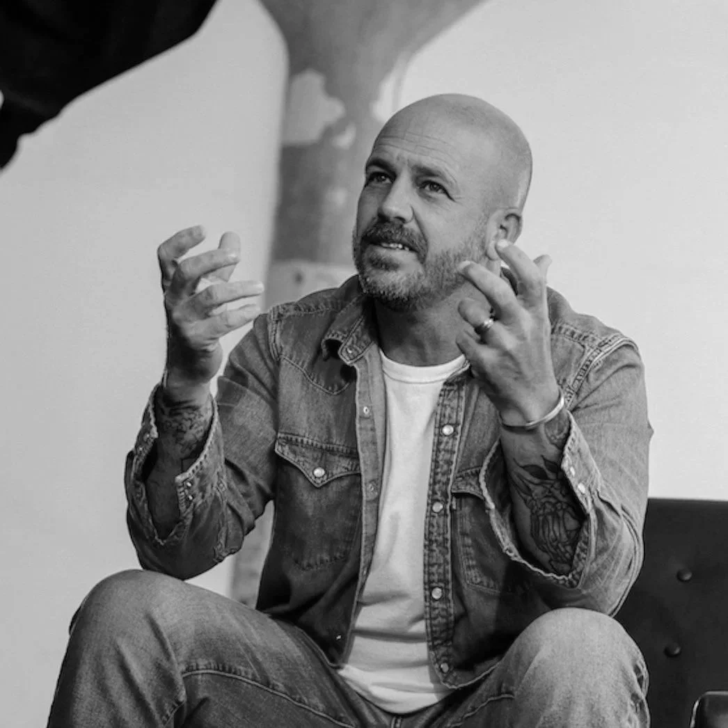 A man with a beard and tattoos wearing a denim jacket and white t-shirt, sitting and gesturing with his hands as if talking or explaining something.