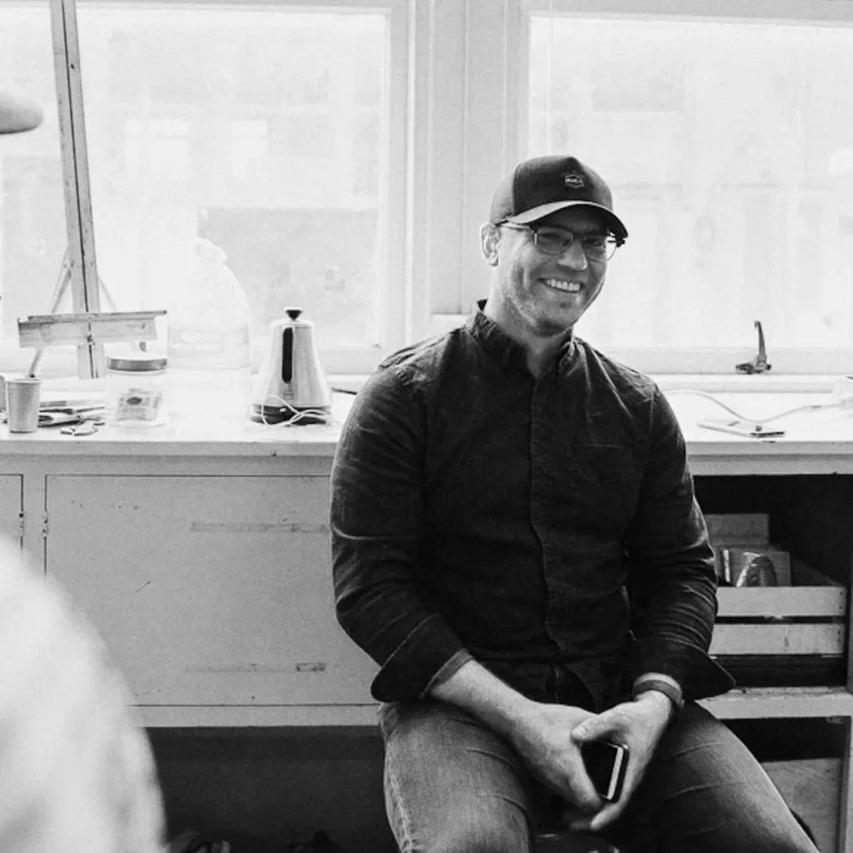 A man in a dark shirt, glasses, and a baseball cap sitting and smiling indoors near a window, holding a phone.