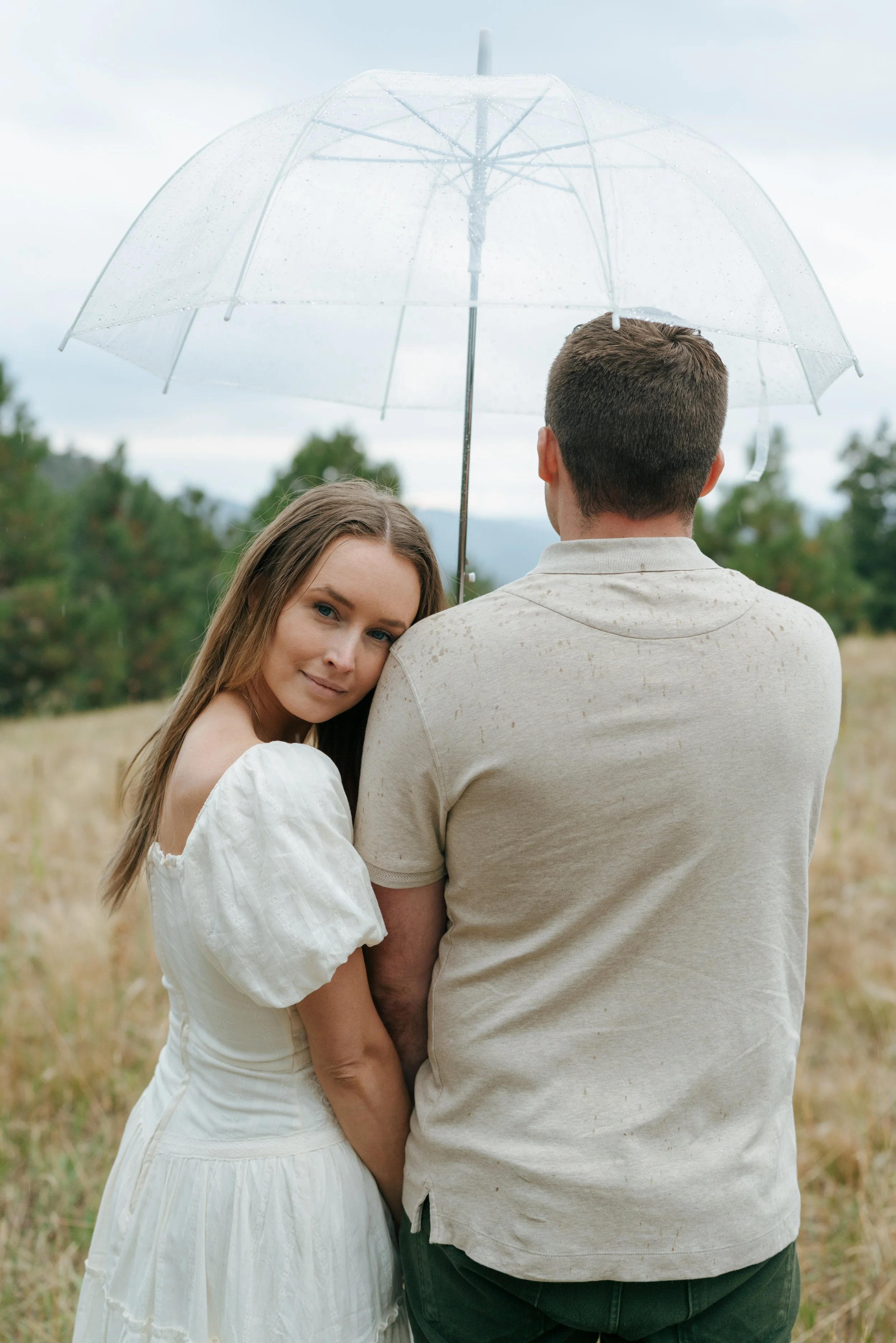 a woman resting her head on a man's shoulder as he holds a clear umbrella
