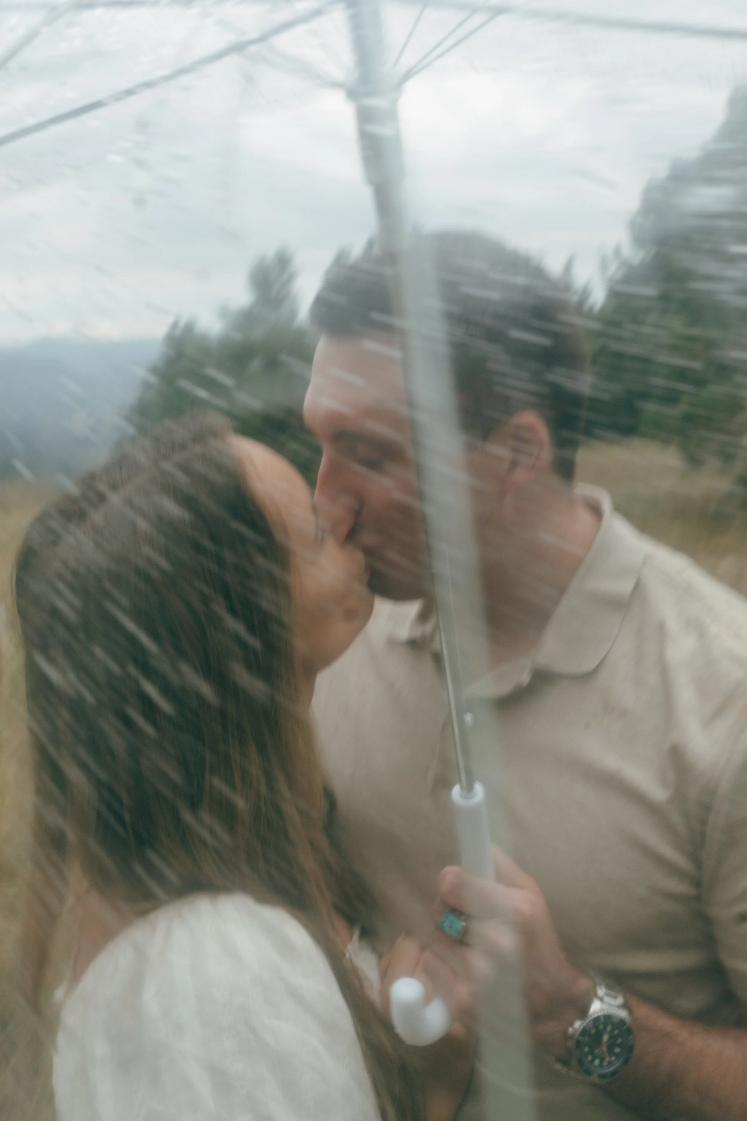 a couple kissing underneath a clear umbrella as it rains during their engagement photoshoot
