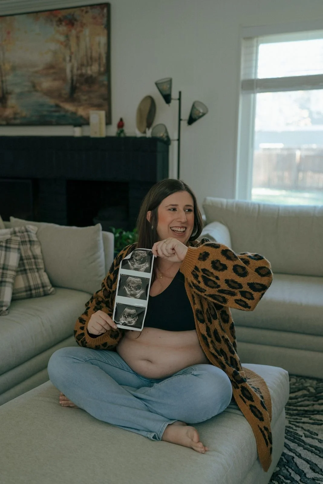 A pregnant woman sits cross-legged on a couch, smiling and holding ultrasound photos in a living room for a half baked pregnancy photoshootx