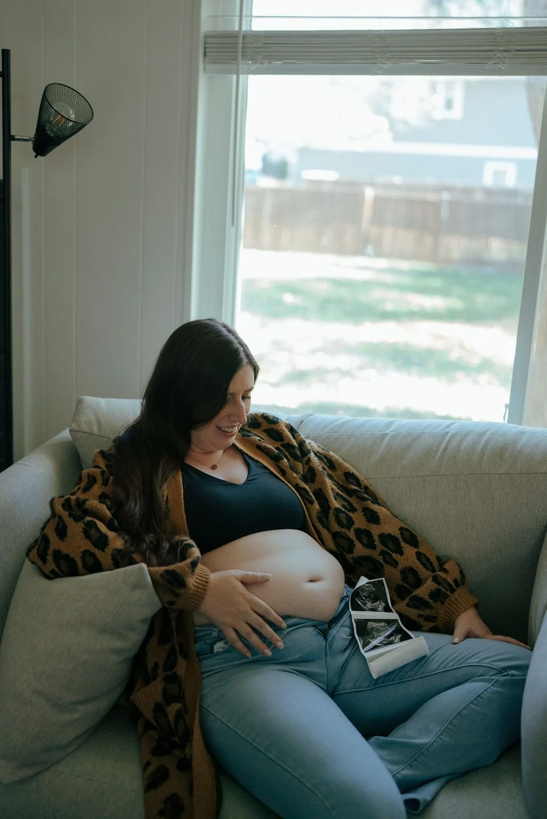 A pregnant woman sits on a couch, touching her bare belly and looking at ultrasound photos resting on her lap for a half baked pregnancy photoshoot