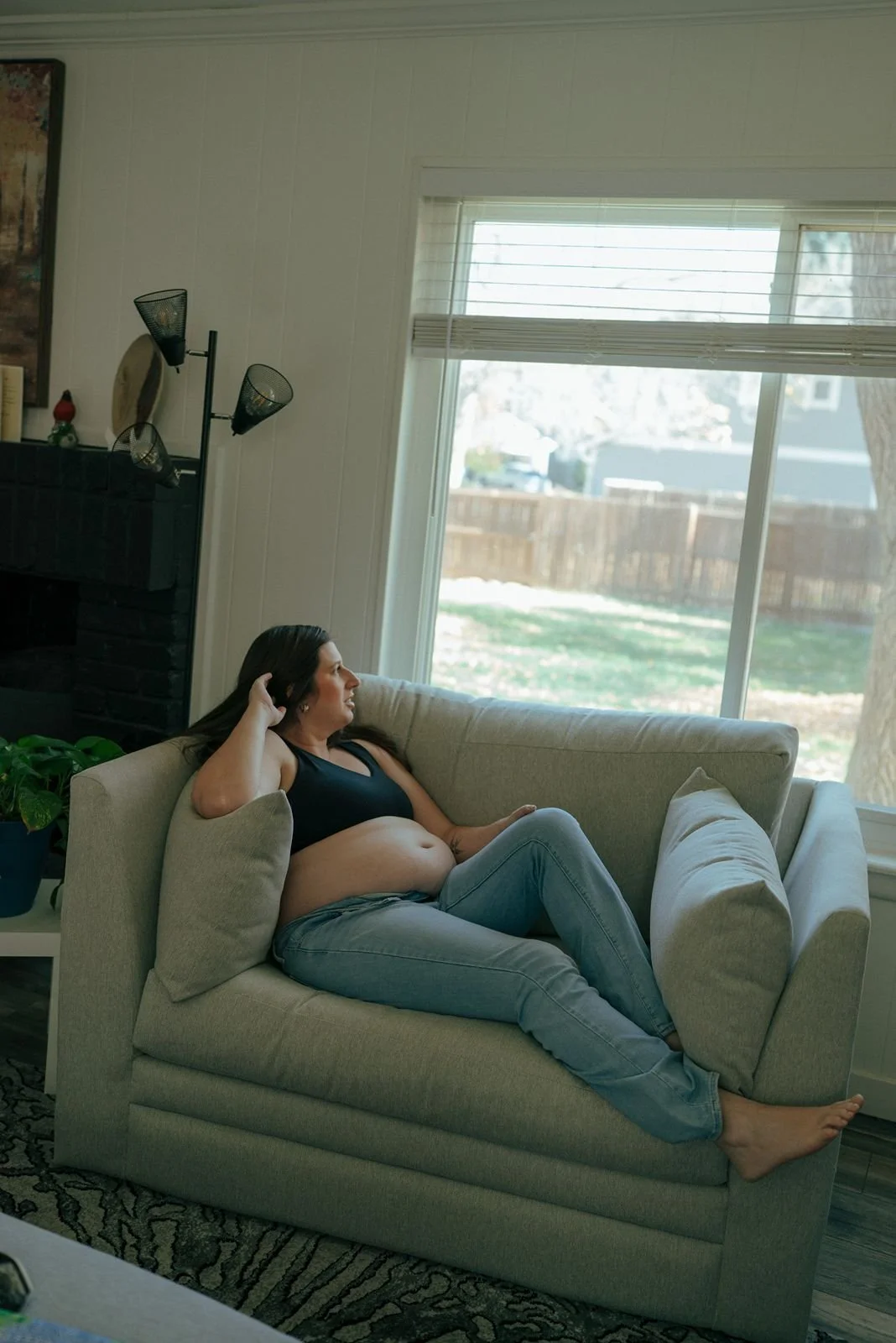 A pregnant woman reclines on a light-colored couch by a window, wearing jeans and a black top, looking outside with one hand resting on her head for a half baked pregnancy photoshoot