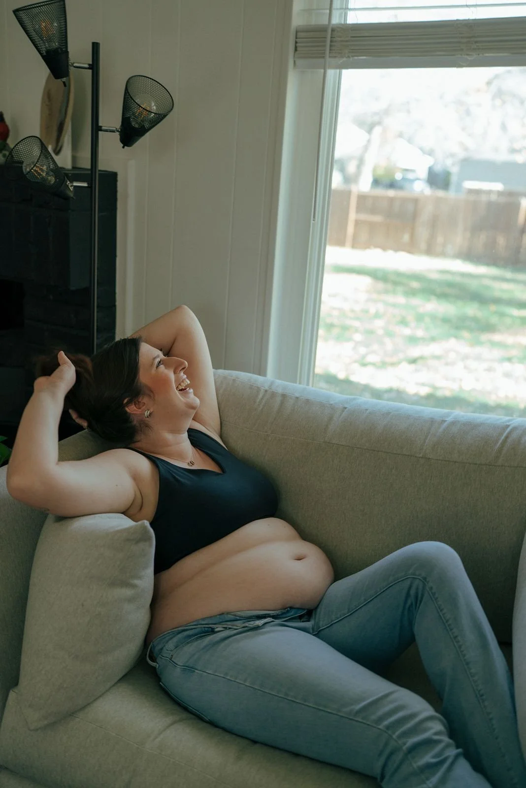 A pregnant woman reclines on a light-colored couch by a window, wearing jeans and a black top, looking outside with one hand resting on her head for a half baked pregnancy photoshoot
