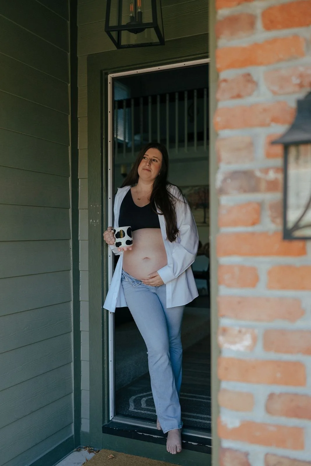 Pregnant woman standing barefoot in a doorway, wearing jeans, a black top, and an unbuttoned white shirt,  for a half baked pregnancy photoshoot