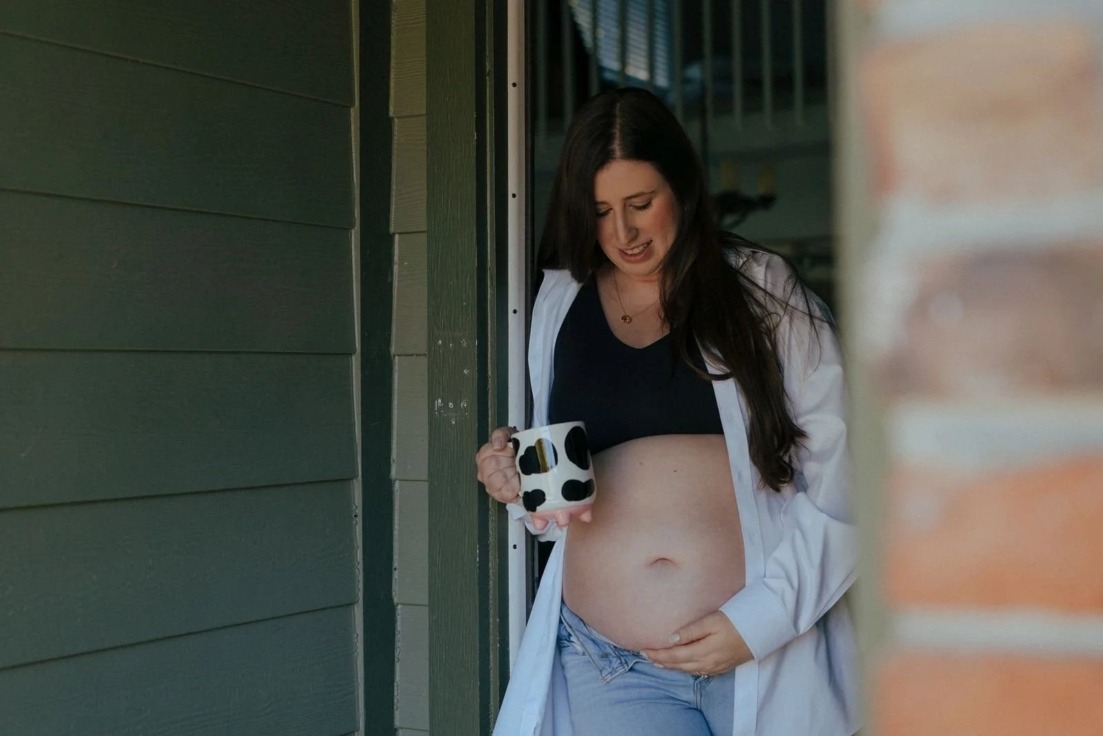 Pregnant woman standing barefoot in a doorway, wearing jeans, a black top, and an unbuttoned white shirt,  for a half baked pregnancy photoshoot