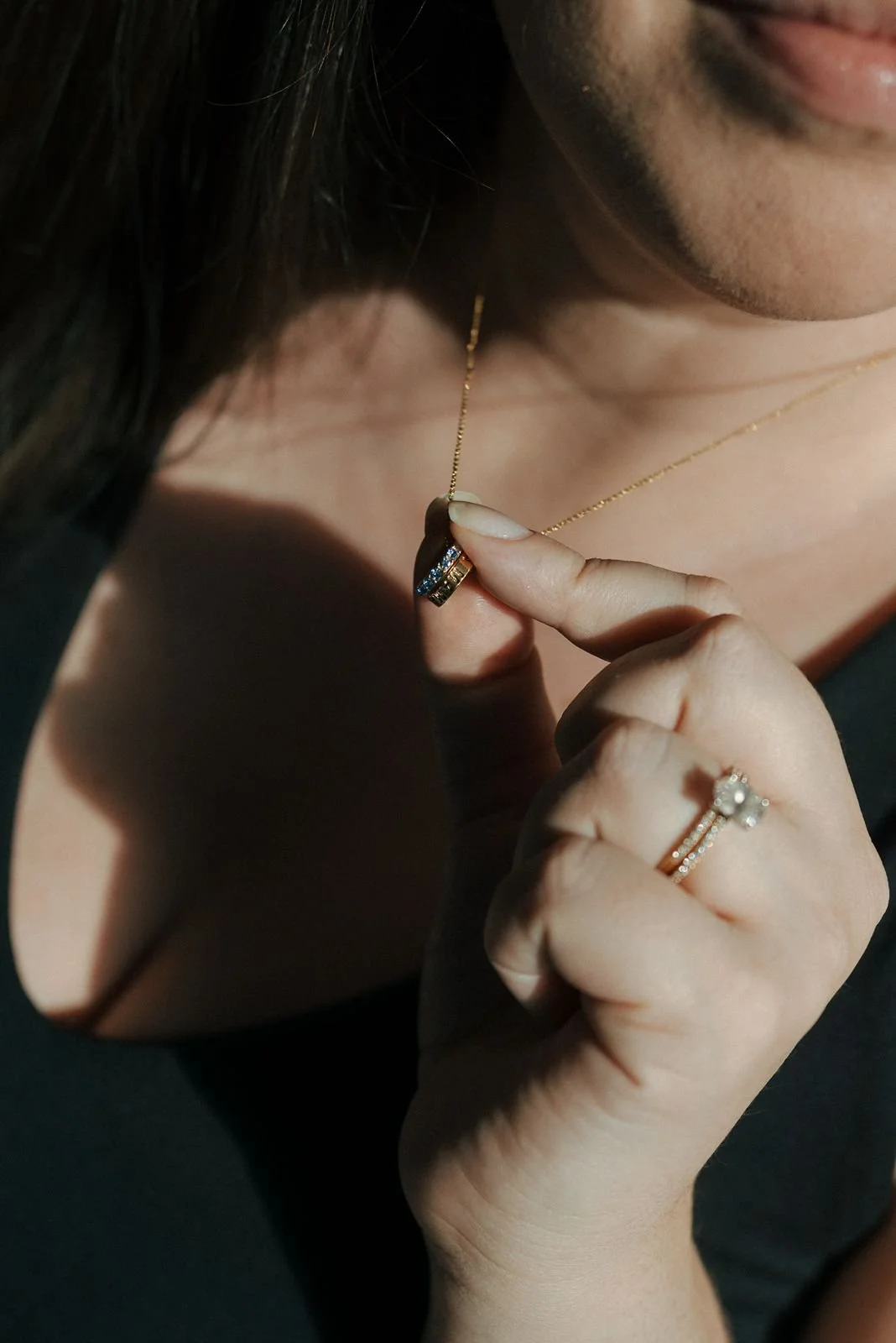 A woman holds a small pendant on a gold necklace; she wears a black top and a gold ring with a flower-shaped design on her finger.
