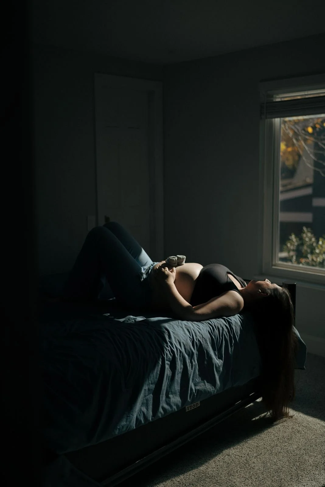 A pregnant woman lies on her back on a bed in a dimly lit room, with light coming through a nearby window for a half baked pregnancy photoshoot
