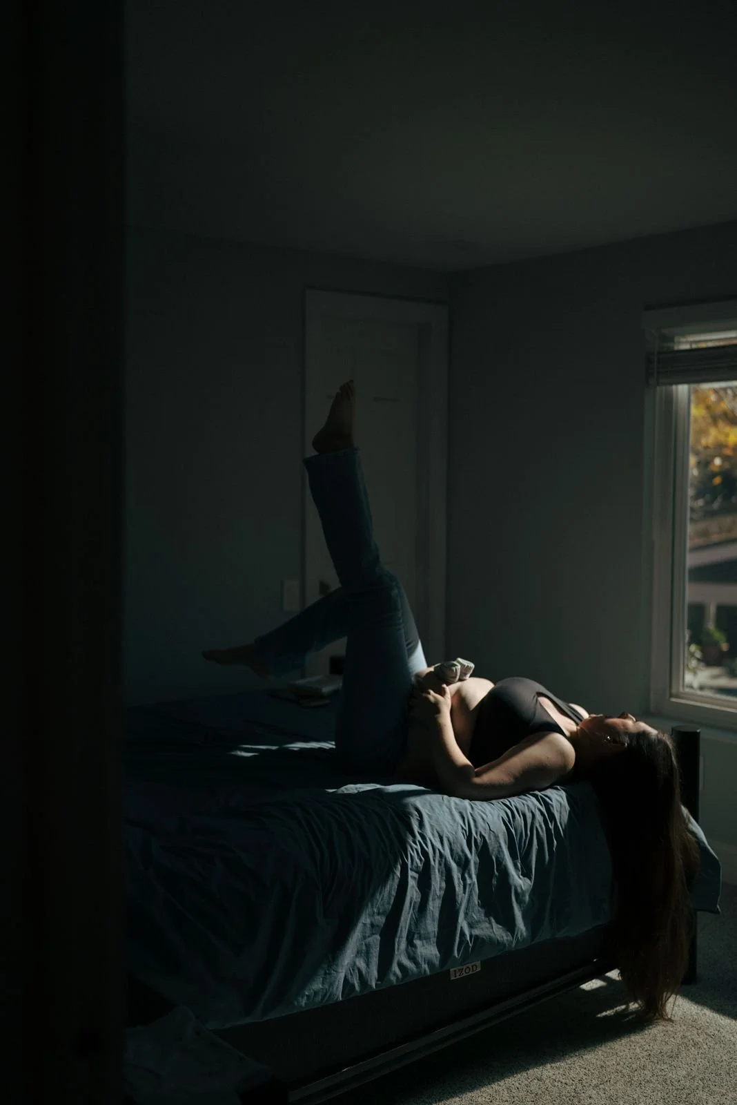 A pregnant woman lies on her back on a bed in a dimly lit room, with light coming through a nearby window for a half baked pregnancy photoshoot