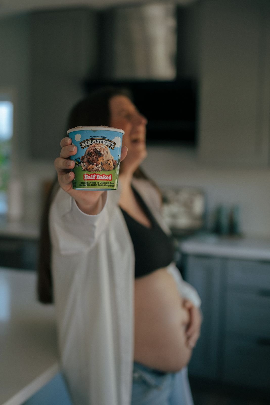 A person in a kitchen holds up a pint of Ben &amp; Jerry's Half Baked ice cream, with their other hand resting on their exposed belly.