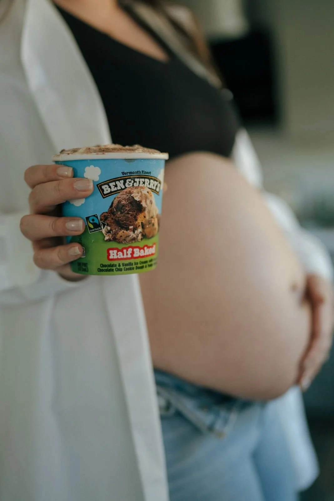 A person in a kitchen holds up a pint of Ben &amp; Jerry's Half Baked ice cream, with their other hand resting on their exposed belly.