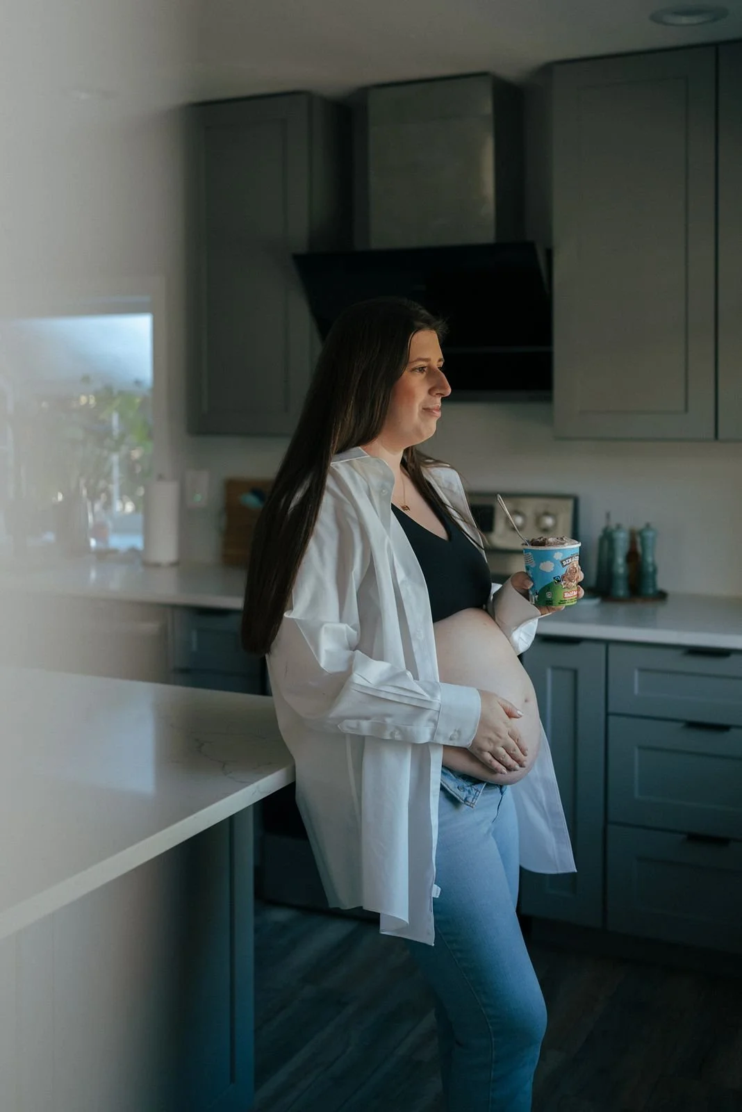 A person with a visible pregnant belly holds a tub of Ben &amp; Jerry's ice cream and a spoon while wearing jeans and an open shirt.