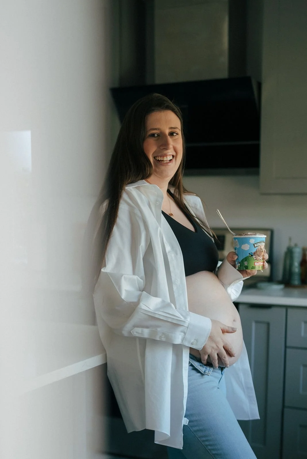 A person with a visible pregnant belly holds a tub of Ben &amp; Jerry's ice cream and a spoon while wearing jeans and an open shirt.