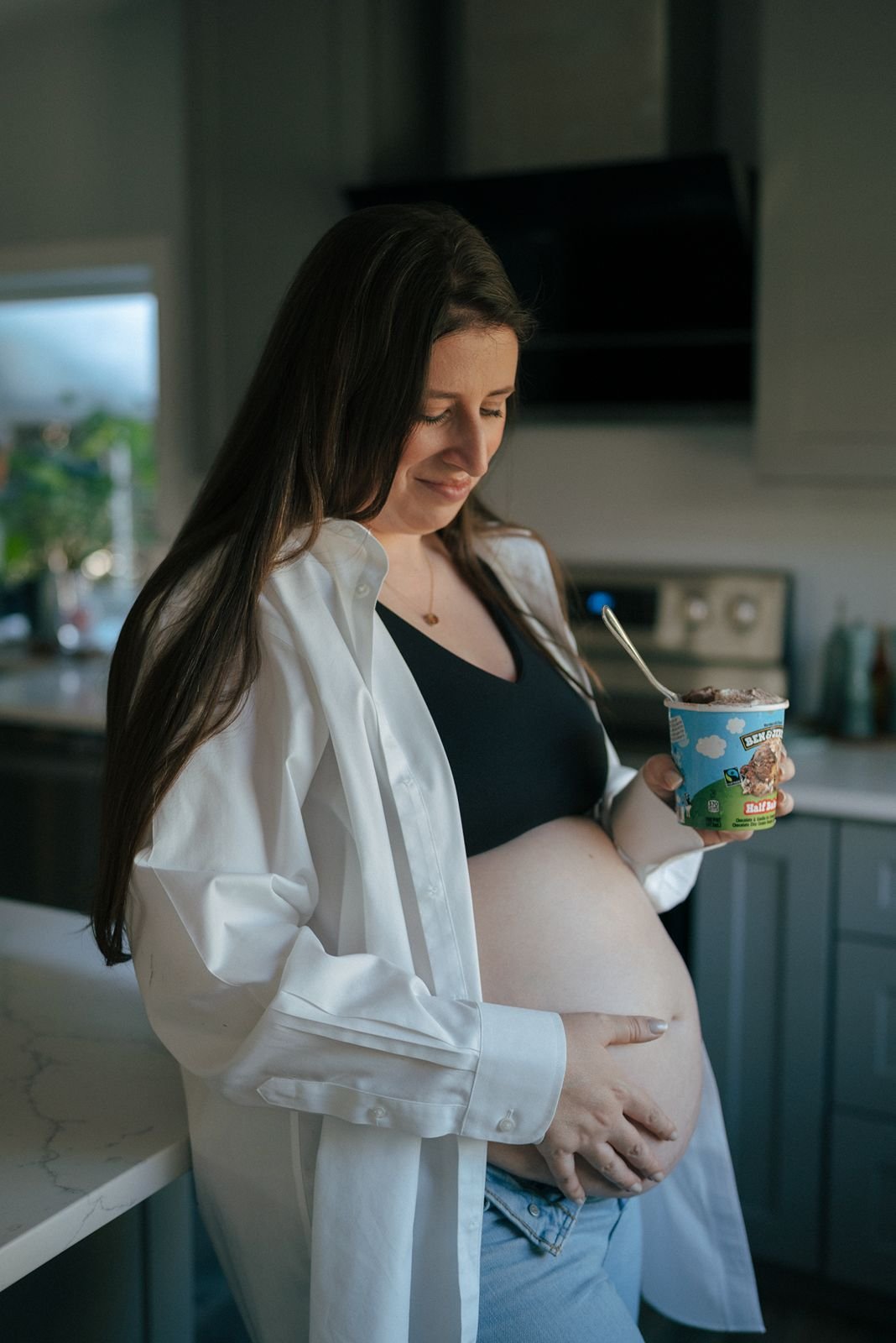 A person with a visible pregnant belly holds a tub of Ben &amp; Jerry's ice cream and a spoon while wearing jeans and an open shirt.