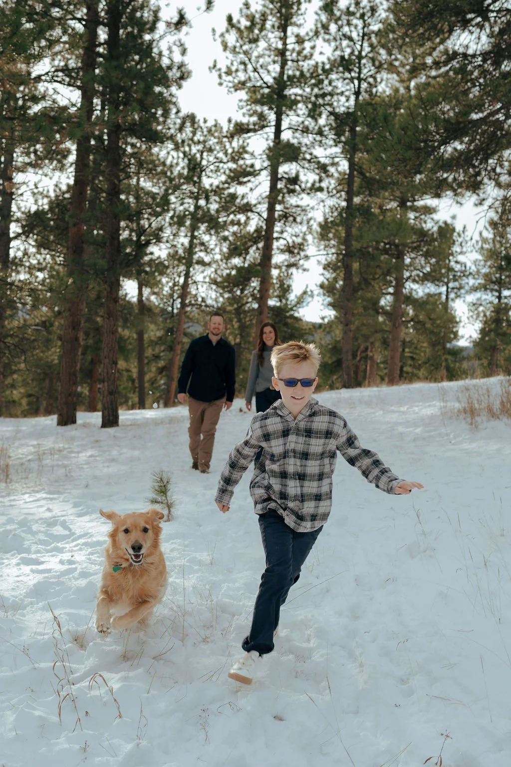 A boy wearing a plaid jacket walks in a snowy forest with a golden retriever for a documentary family photography session