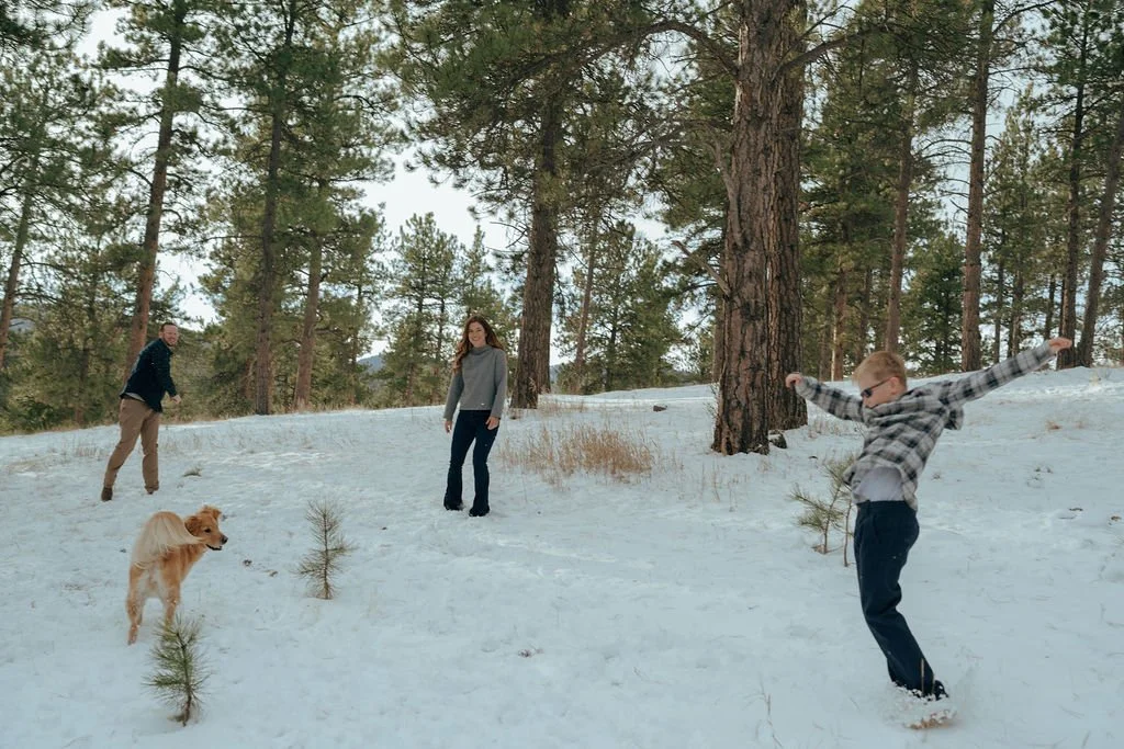 Three people and a dog are playing in a snowy forested area for a documentary family photography  session
