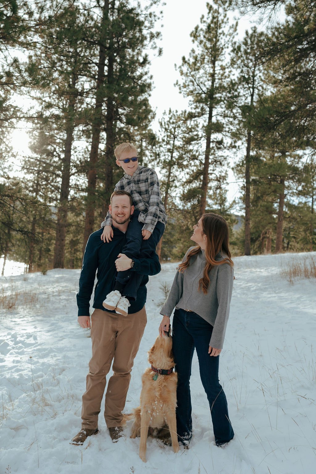 A man, woman, child, and a golden retriever stand together in a snowy forest with pine trees in the background for a documentary family photography session