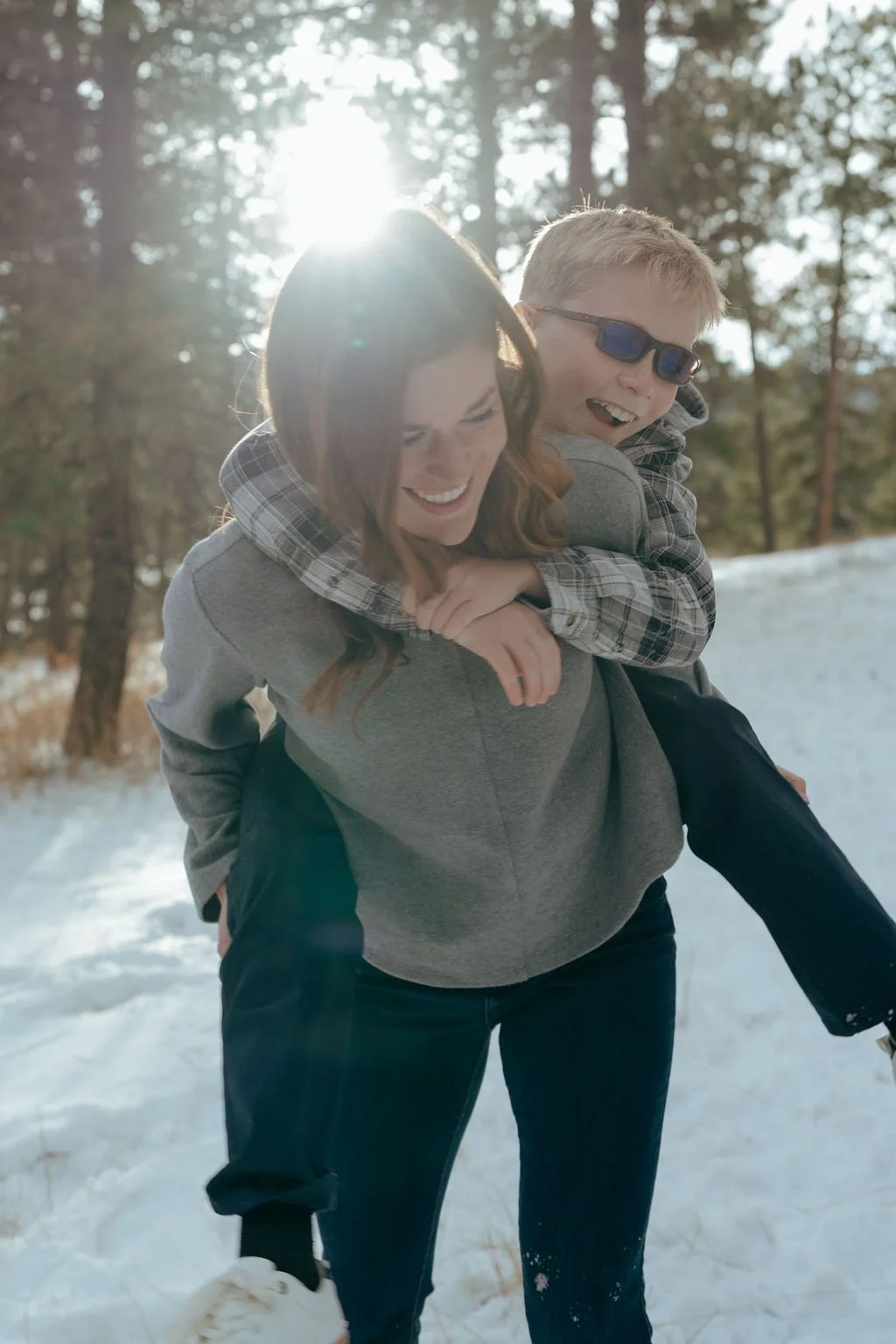Woman carrying a smiling boy in sunglasses on her back in a snowy forest with sunlight shining through the trees.