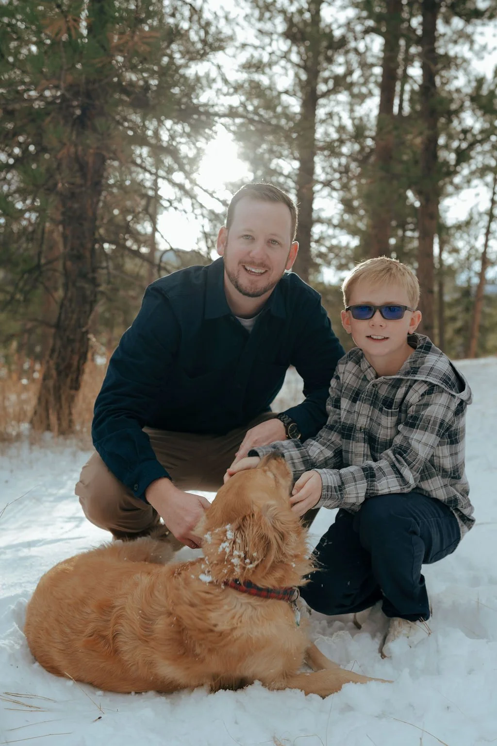 A man and a boy kneel in the snow petting a golden retriever, with a forest of pine trees in the background.