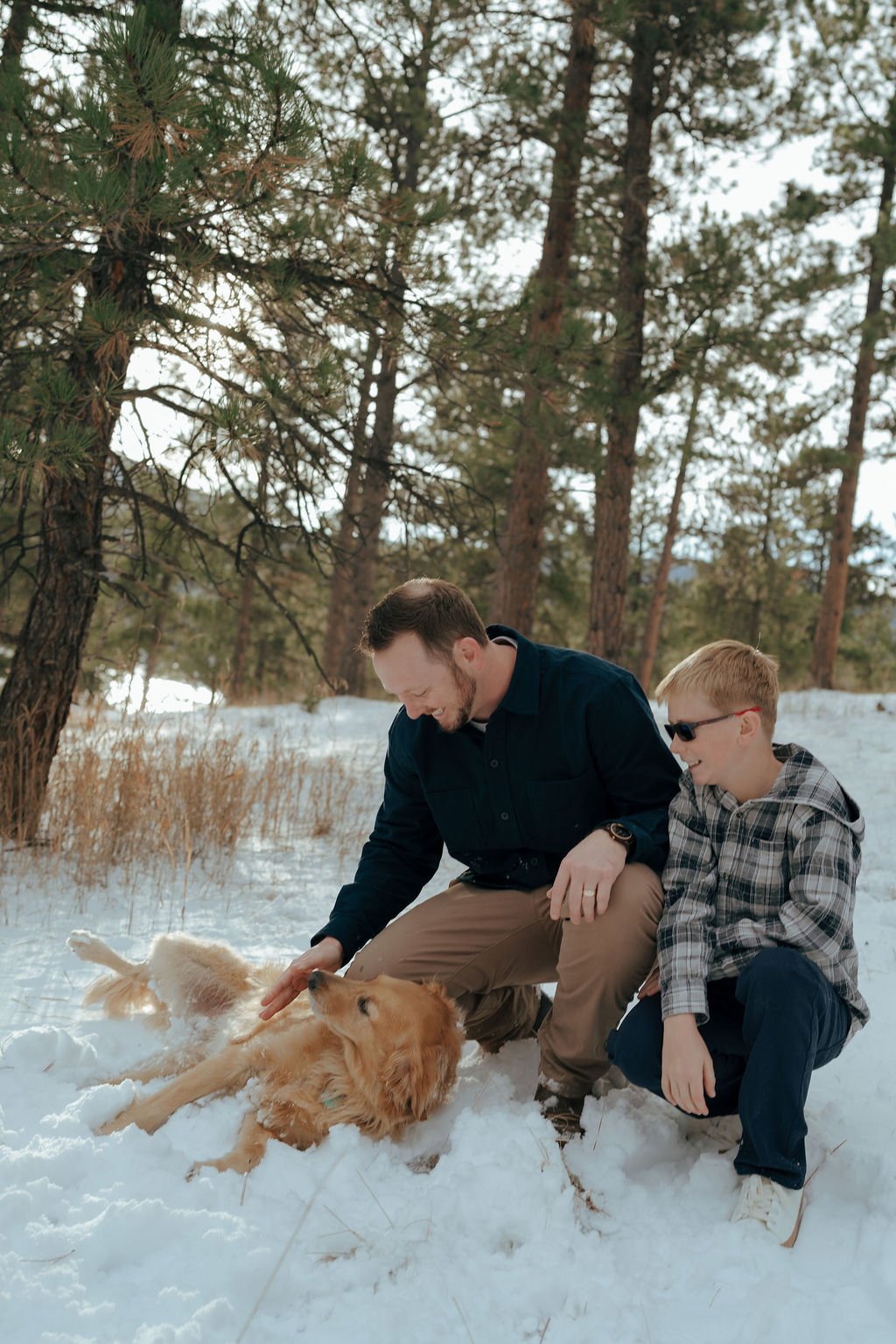 A man and a boy kneel in the snow petting a golden retriever, with a forest of pine trees in the background.