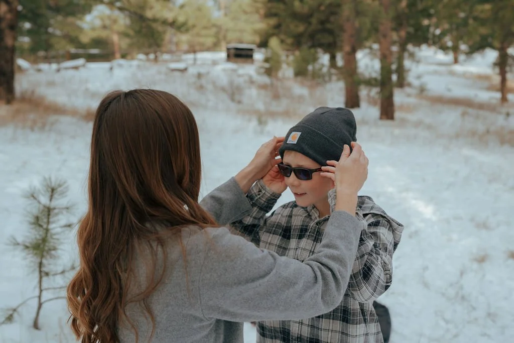 An adult adjusts a beanie on a child wearing sunglasses in a snowy, wooded outdoor setting.