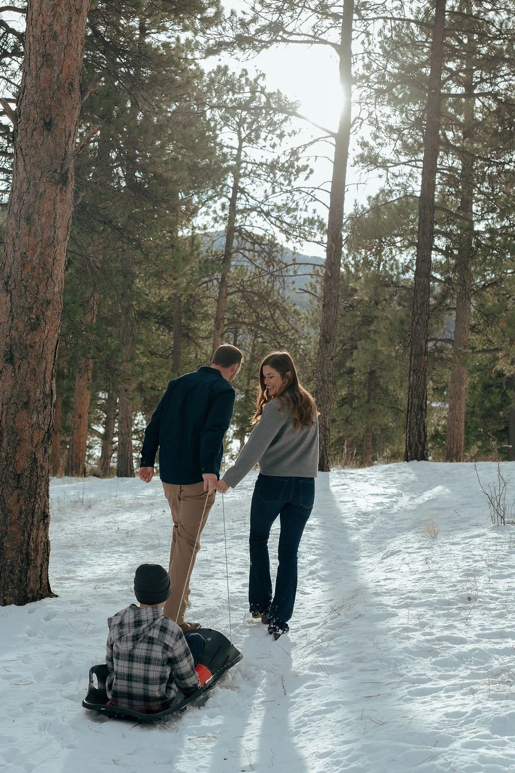 Two adults pull a child in a sled through a snowy forest with tall pine trees, sunlight shining through the branches.