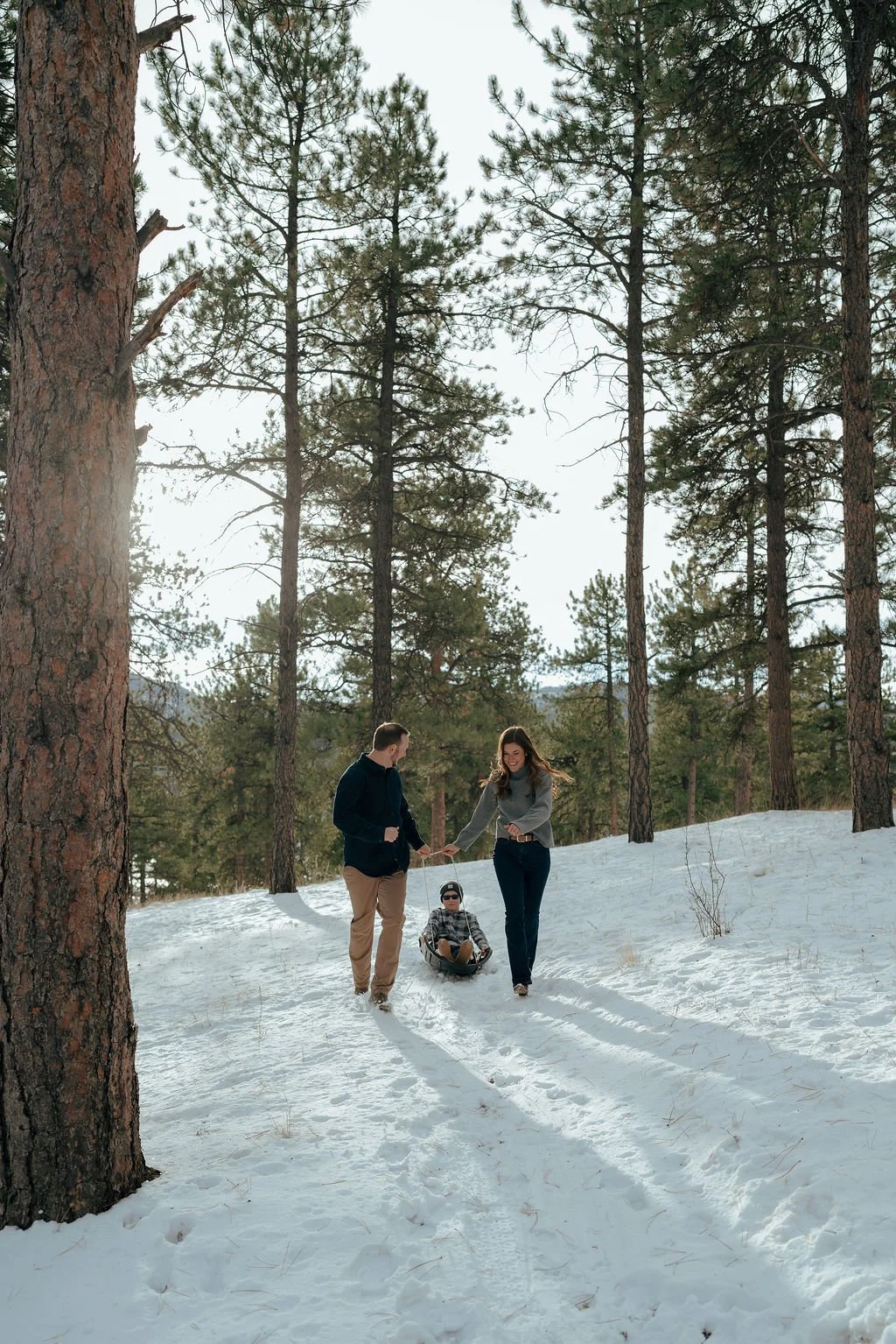 A man and woman walk on a snowy path in a forest while a child rides a sled behind them.